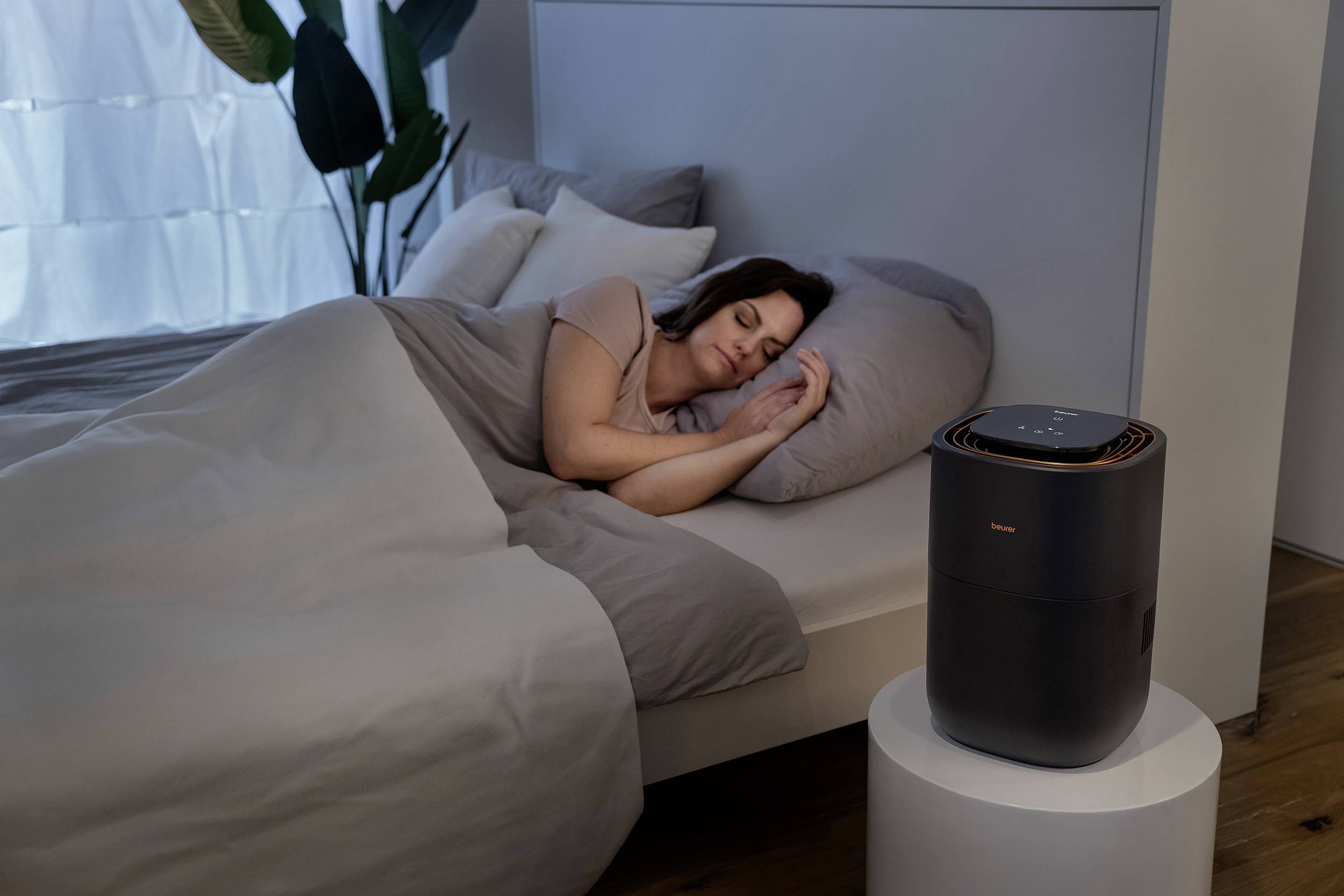 A person is sleeping in a bed in a bedroom. An air purifier is standing on a table next to the bed.
