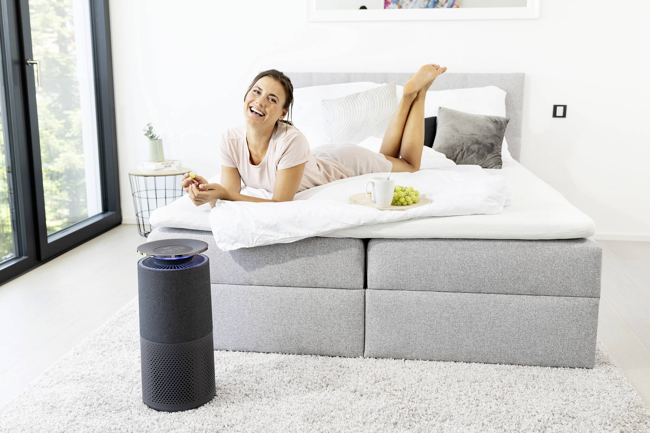 A smiling person is lying relaxed on a bed, eating grapes, while an air purifier stands next to the bed.