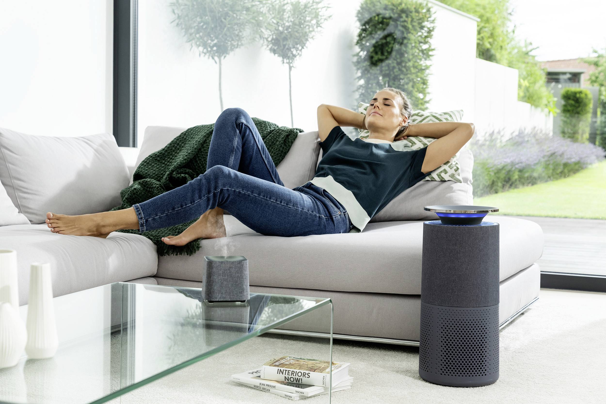 A woman relaxes on a sofa with a speaker in the foreground, in a modern, light-filled living room.