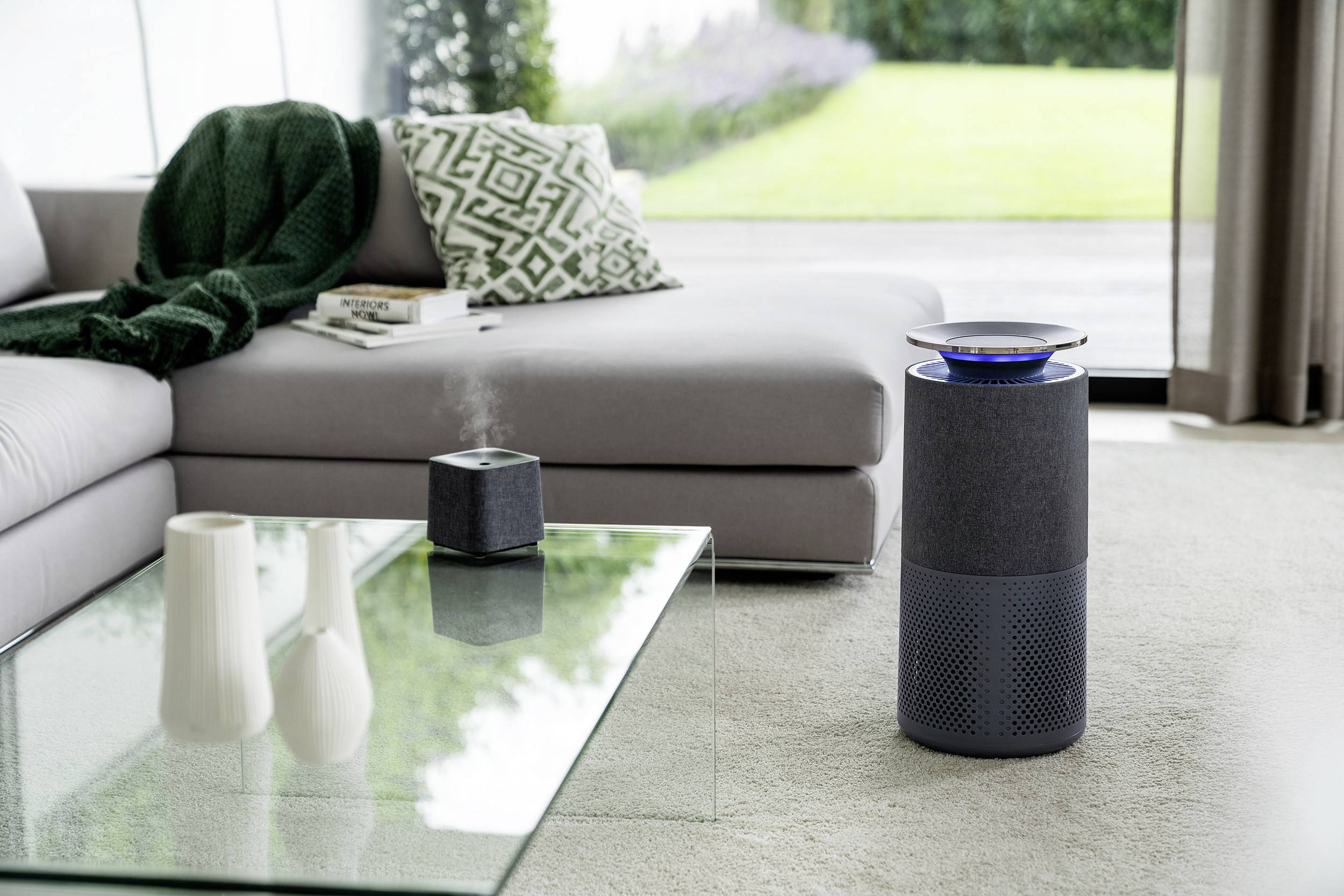 In a modern living room, an air purifier stands on the floor. Another small device sits on a glass table next to a sofa.