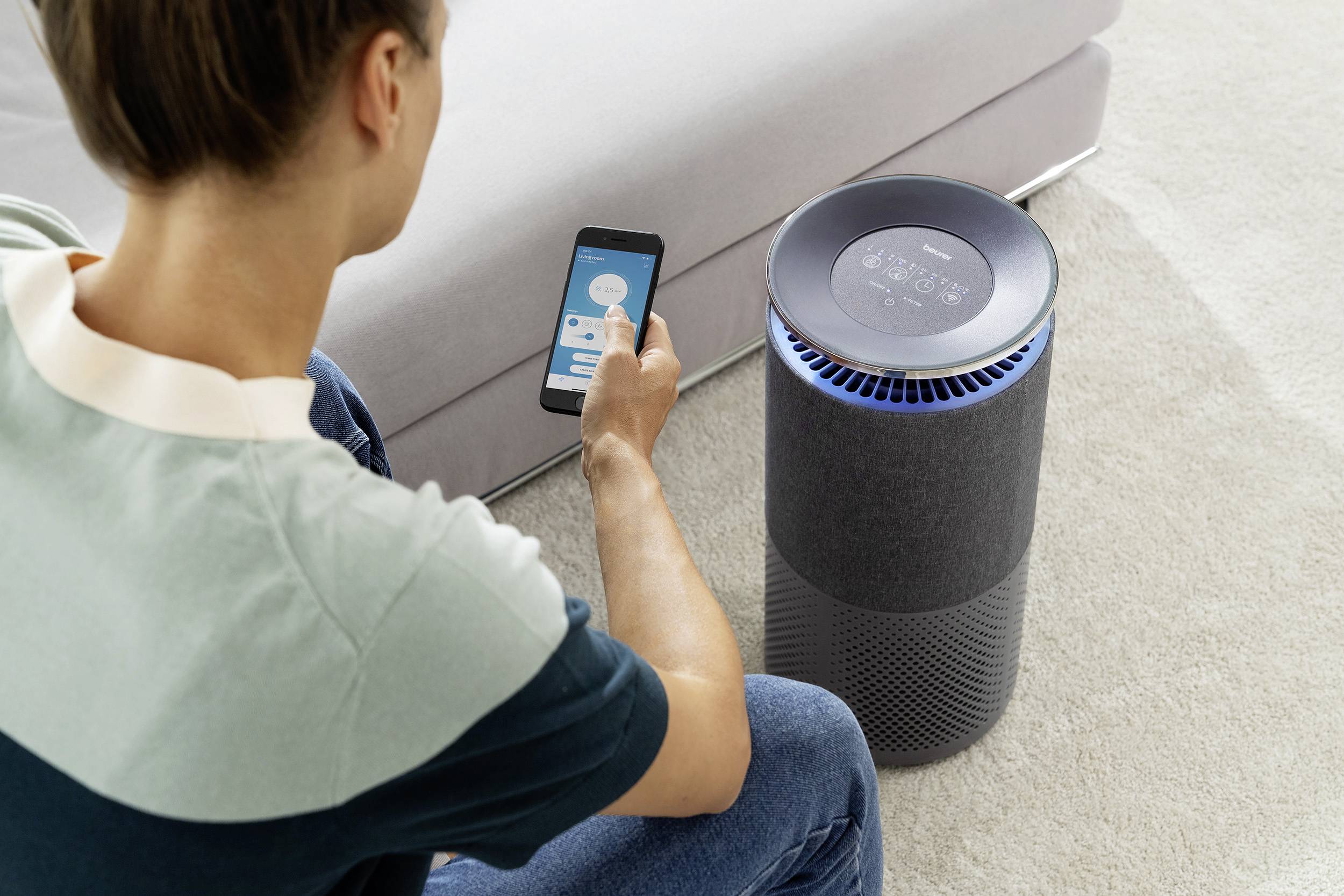 A person is using an app on a mobile phone to control an air purifier next to a sofa. The air purifier is switched on.