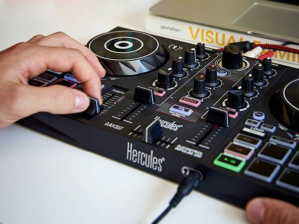 A person is operating a DJ mixing desk with multiple sliders and buttons. In the background, a book with the title 'VISUAL THINKING' is lying down.