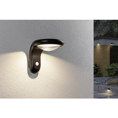 Outdoor wall light with integrated motion sensor, switched on, illuminating a concrete wall. Garden area visible in the background.