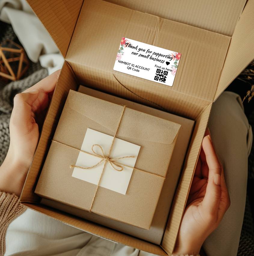 A person is holding an open cardboard box containing a smaller gift package. A sticker on the outside reads 'Thank you for supporting our small business'.