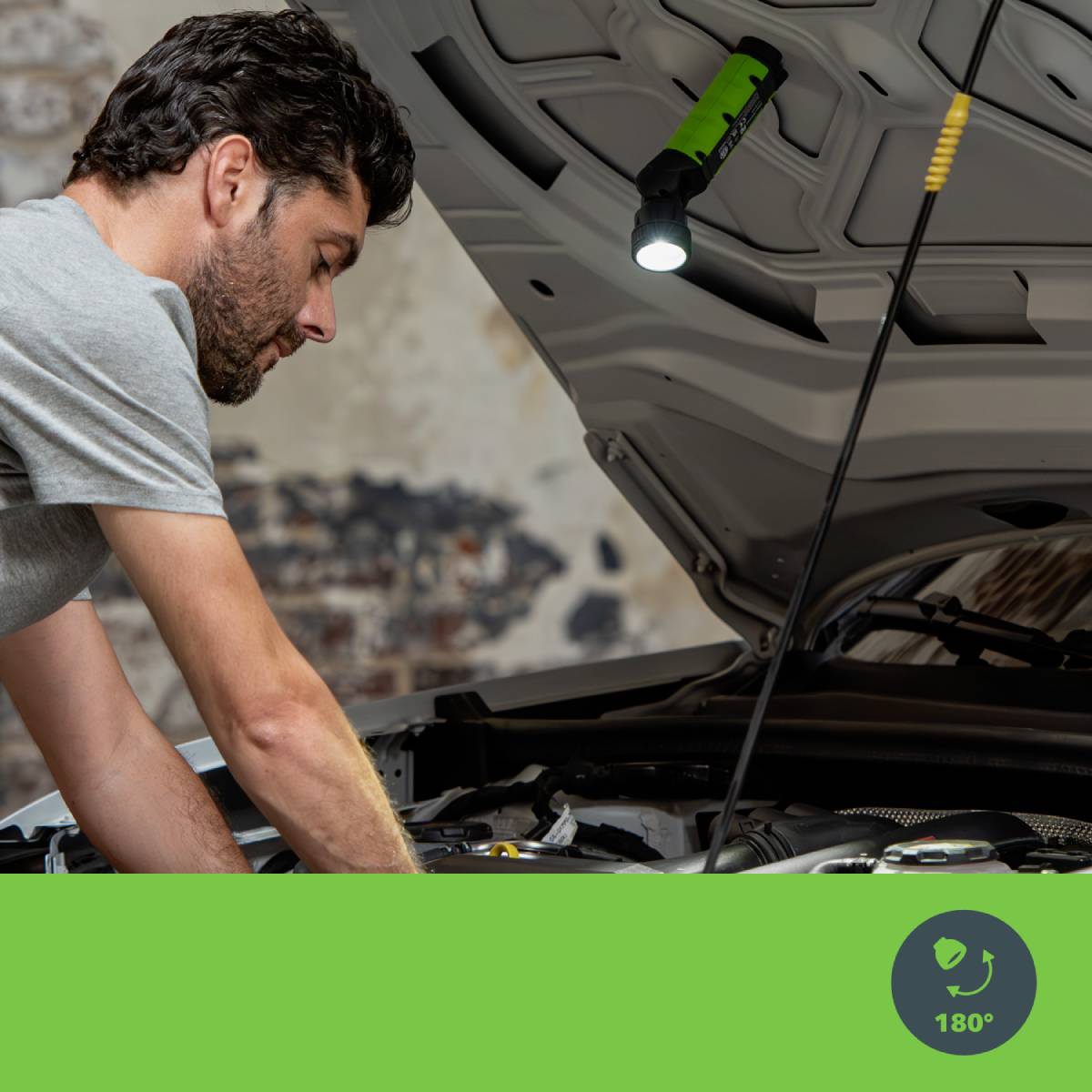 A man examines a car's engine in a garage with the bonnet open. A torch illuminates the work area.