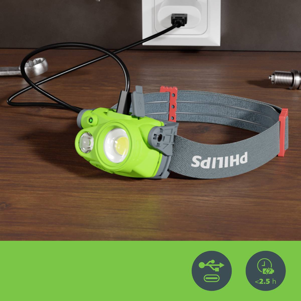 A green head torch with an elastic strap is lying on a table. Plug sockets are visible in the background. Symbolism for rechargeability and an operating time of less than 2.5 hours is apparent.