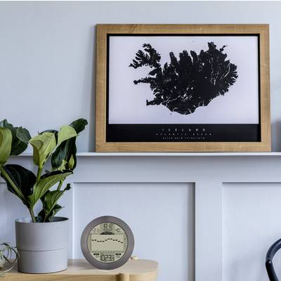 A picture frame with a stylised black outline of an island hangs on a grey wall. Next to it, a plant sits on a shelf.