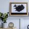 A picture frame with a stylised black outline of an island hangs on a grey wall. Next to it, a plant sits on a shelf.