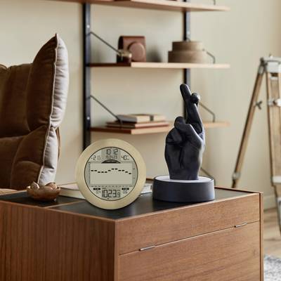 A beige digital alarm clock with time and temperature display sits on a wooden piece of furniture next to a sculpture of a hand in a peace gesture.