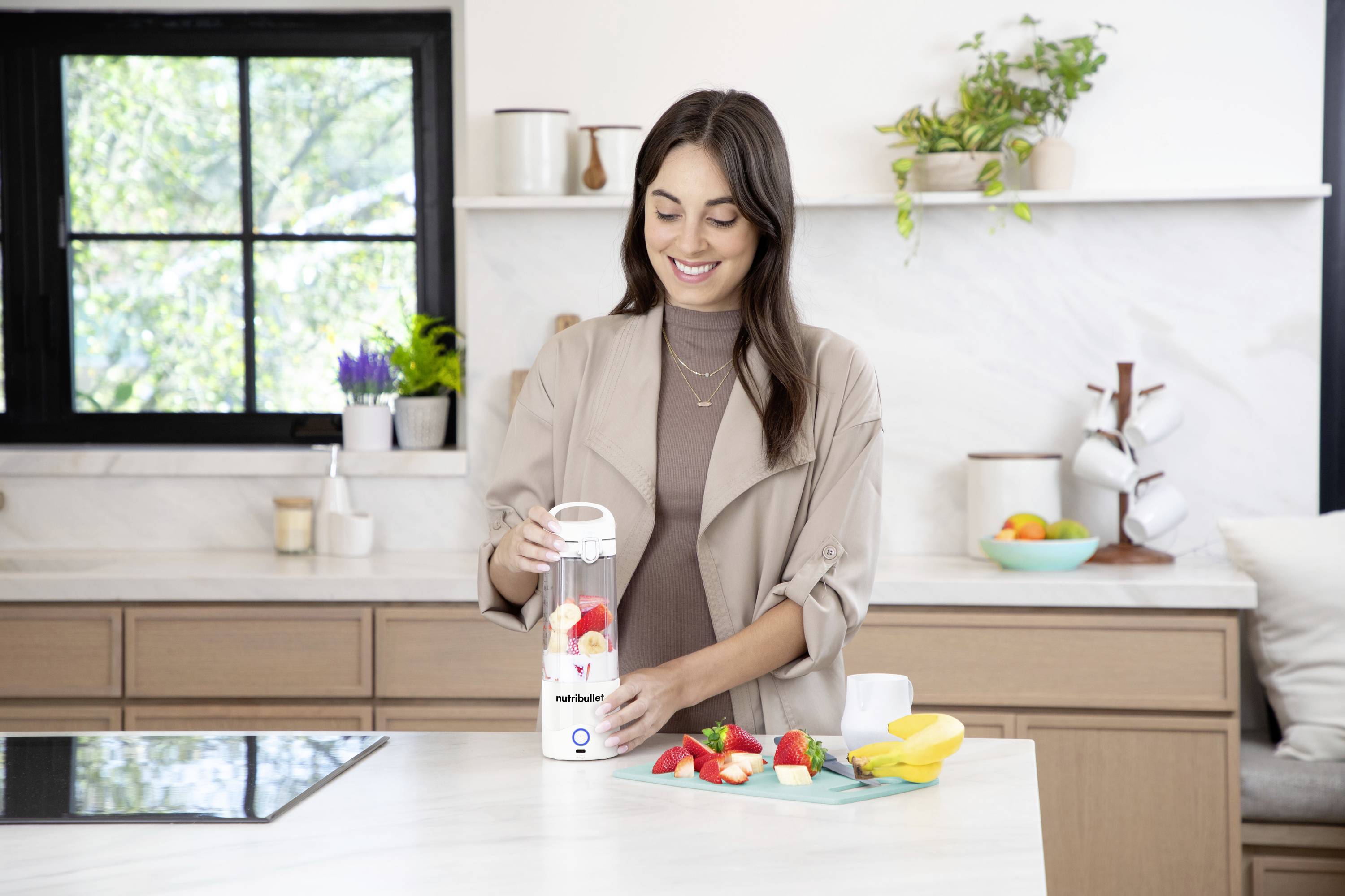 A woman in a modern kitchen is using a hand blender to blend fruits such as strawberries and bananas. She looks content.