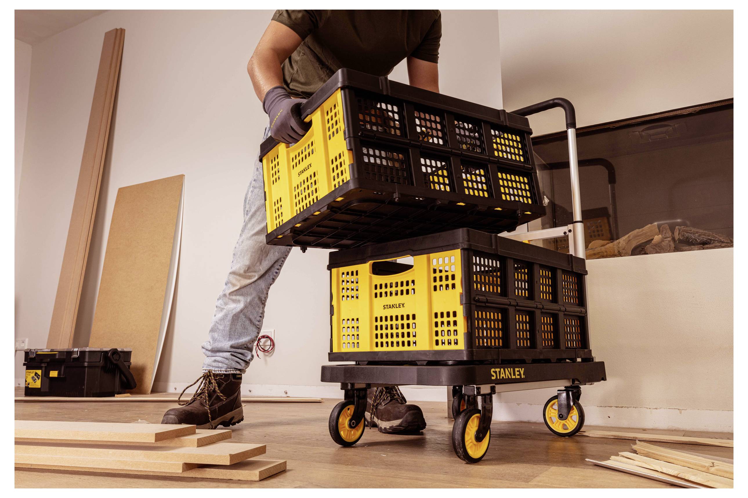 'A person loads yellow and black crates onto a wheeled cart in a workshop or storage area, with wooden planks on the floor.'