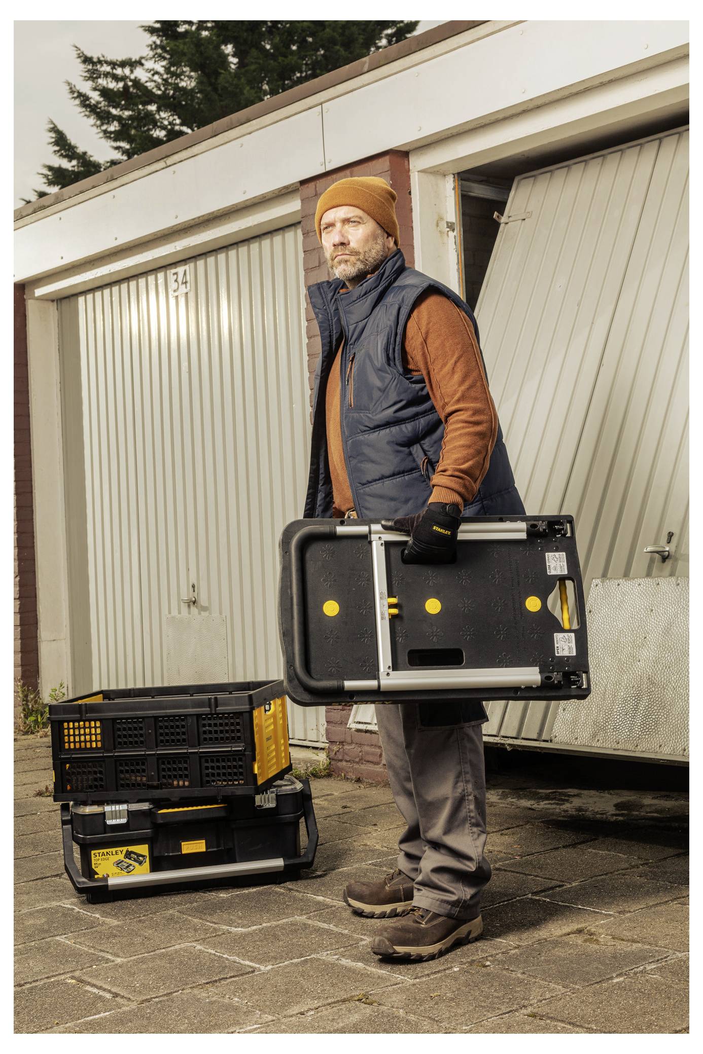 A person stands in front of a garage holding a toolbox, dressed warmly with a hat, gloves, and vest, suggesting they are prepared for work.