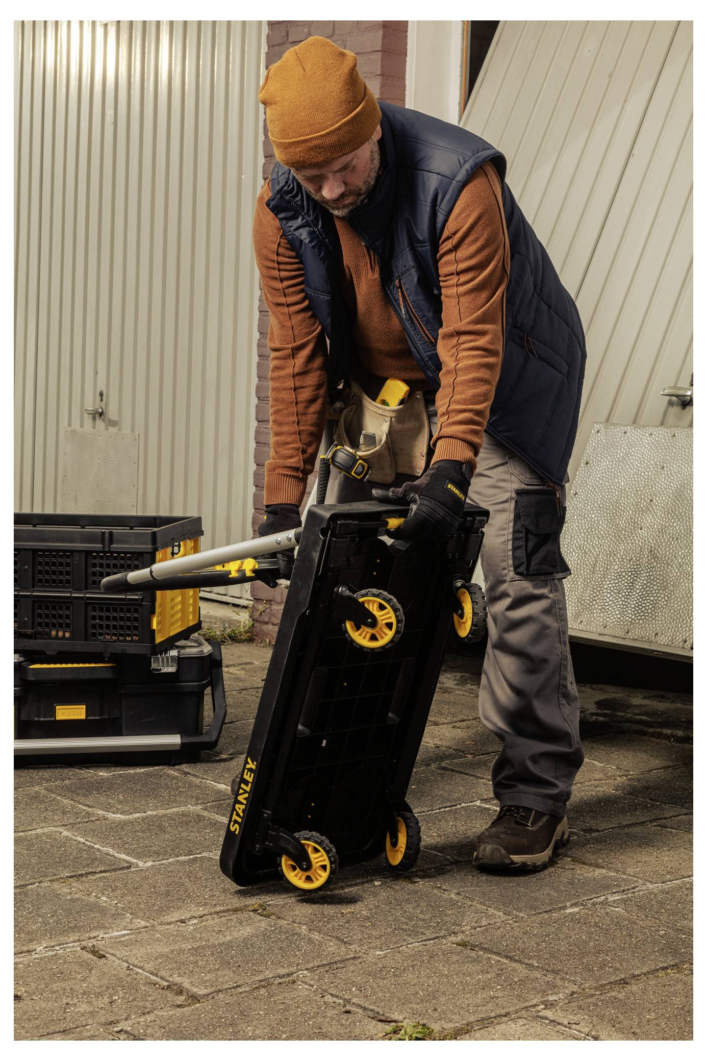 A person in a beanie and vest adjusts a foldable tool trolley with yellow wheels on a paved surface near a garage.