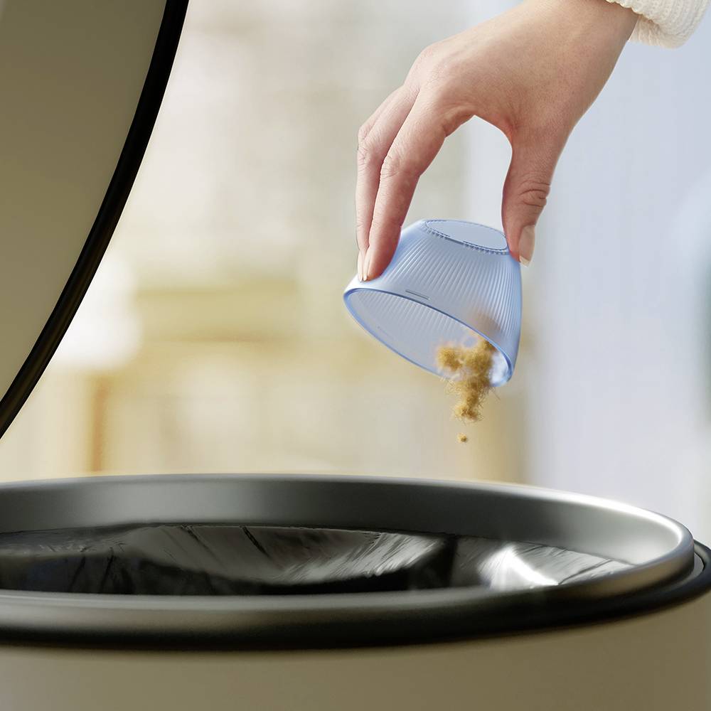 A hand empties crumbs from a dustpan into an open rubbish bin.