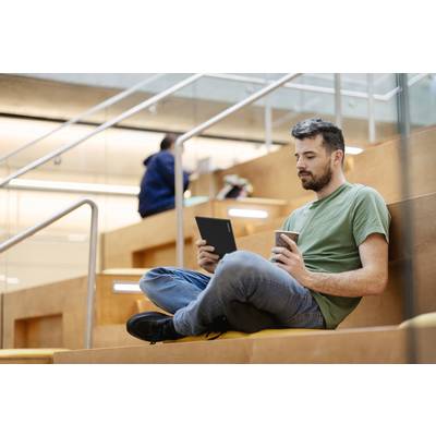 A man is sitting relaxed on a staircase in a modern building, holding a tablet and drinking coffee. Another person is in the background.