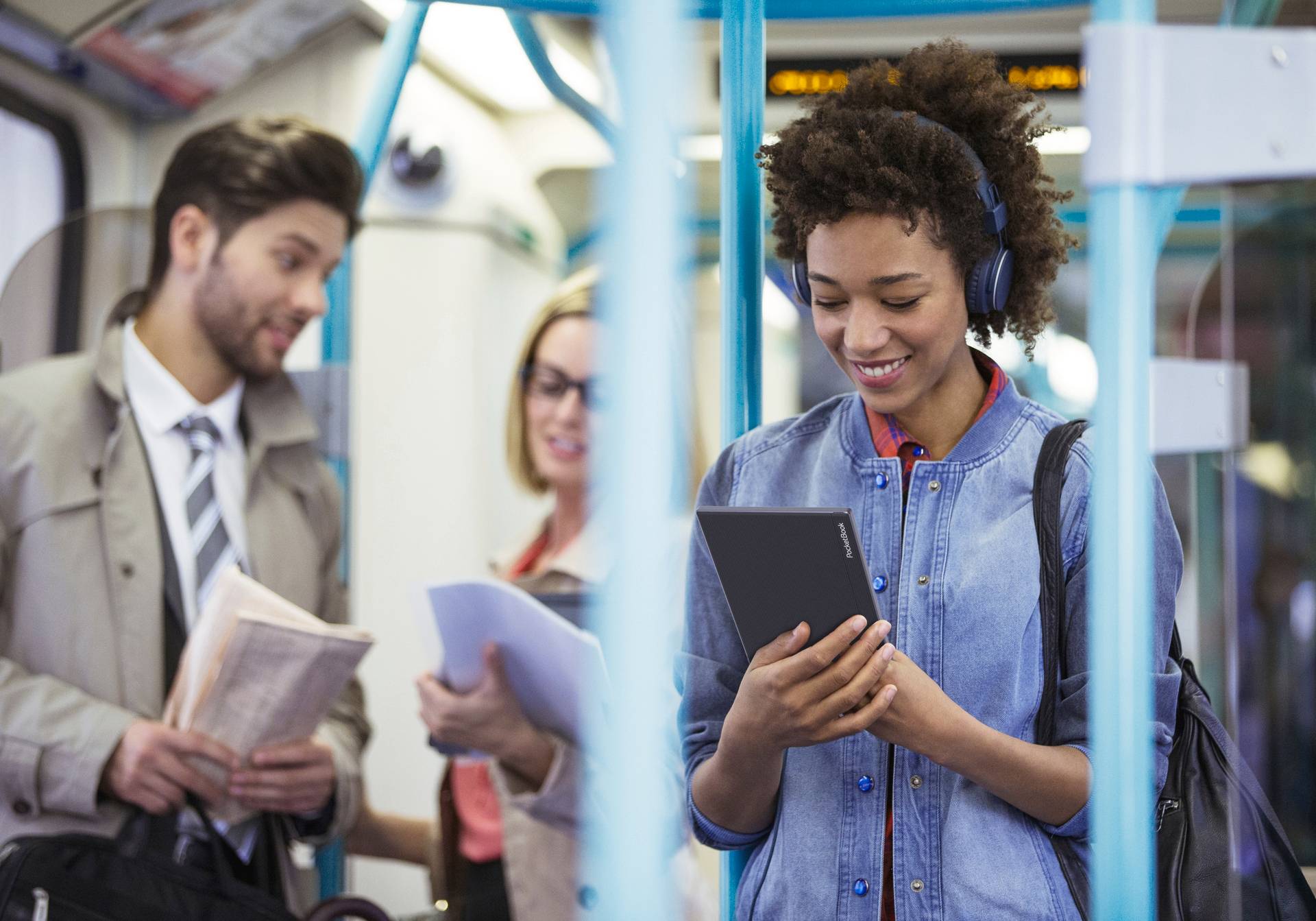 A woman is smiling while reading on a tablet on a train. She is wearing headphones. Two other people are chatting behind her.