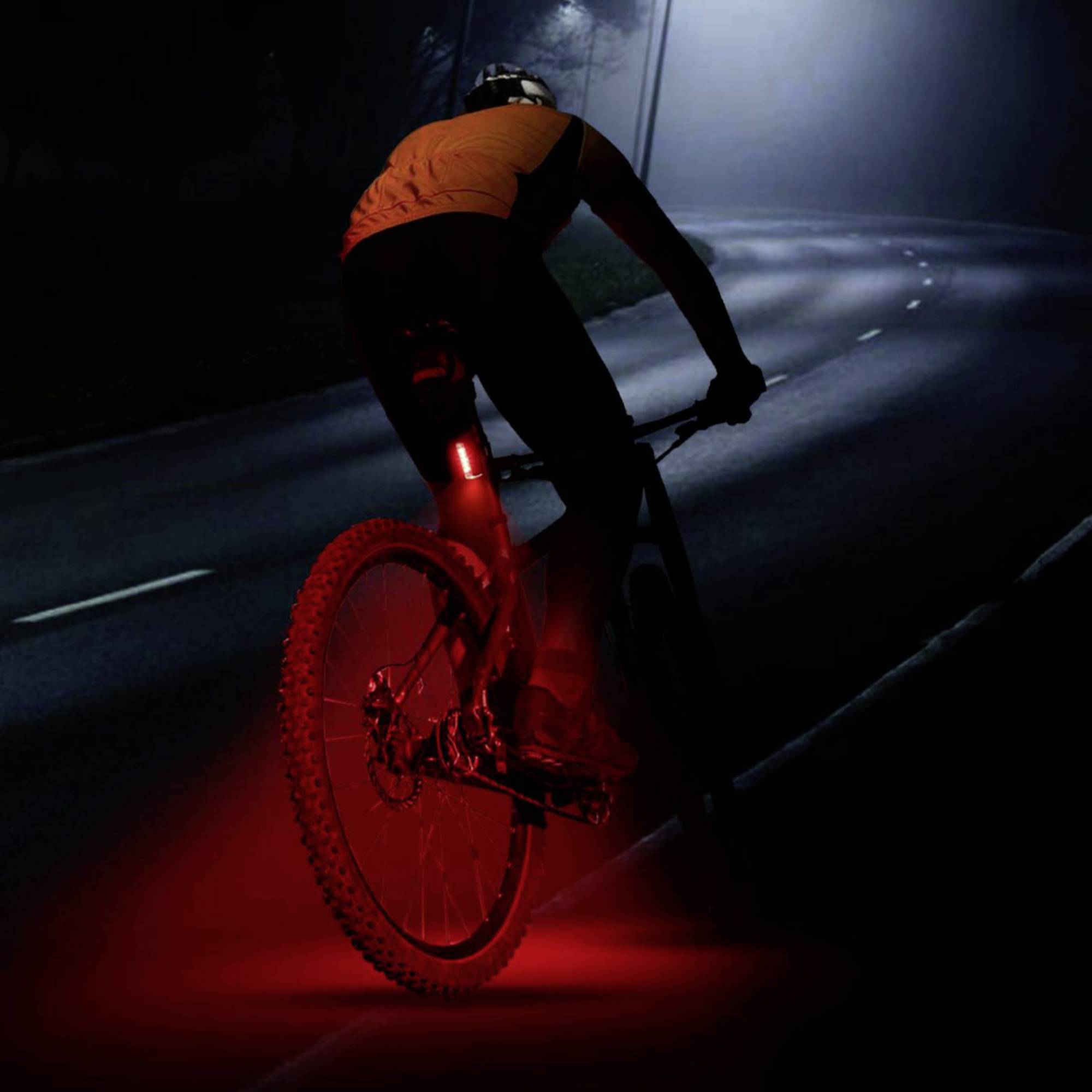 A cyclist with a red rear light is riding at night on an empty street. The headlight illuminates the path ahead of him.