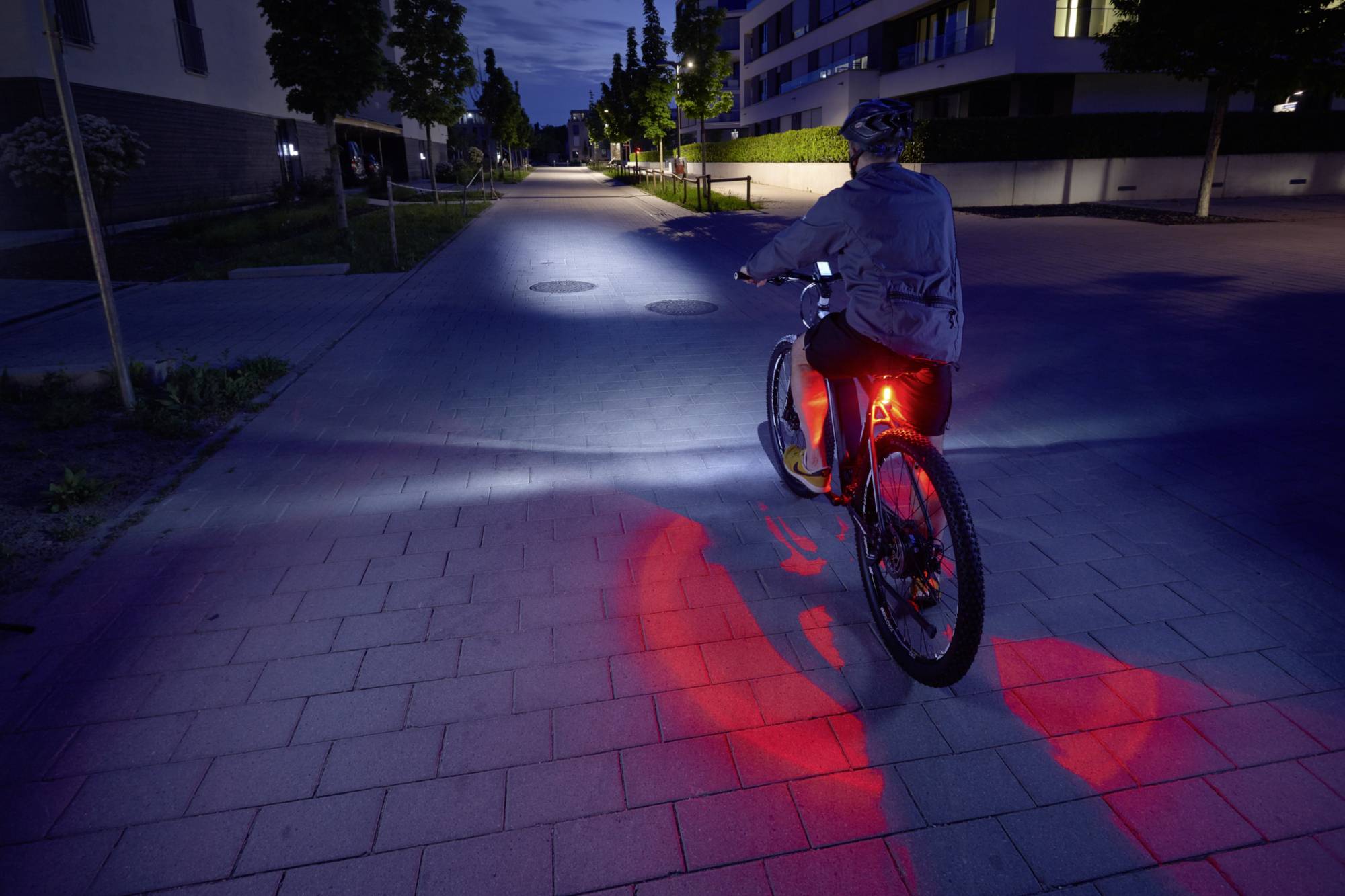 A person is cycling along a lit path at dusk. The bicycle has a red rear light and a headlight.