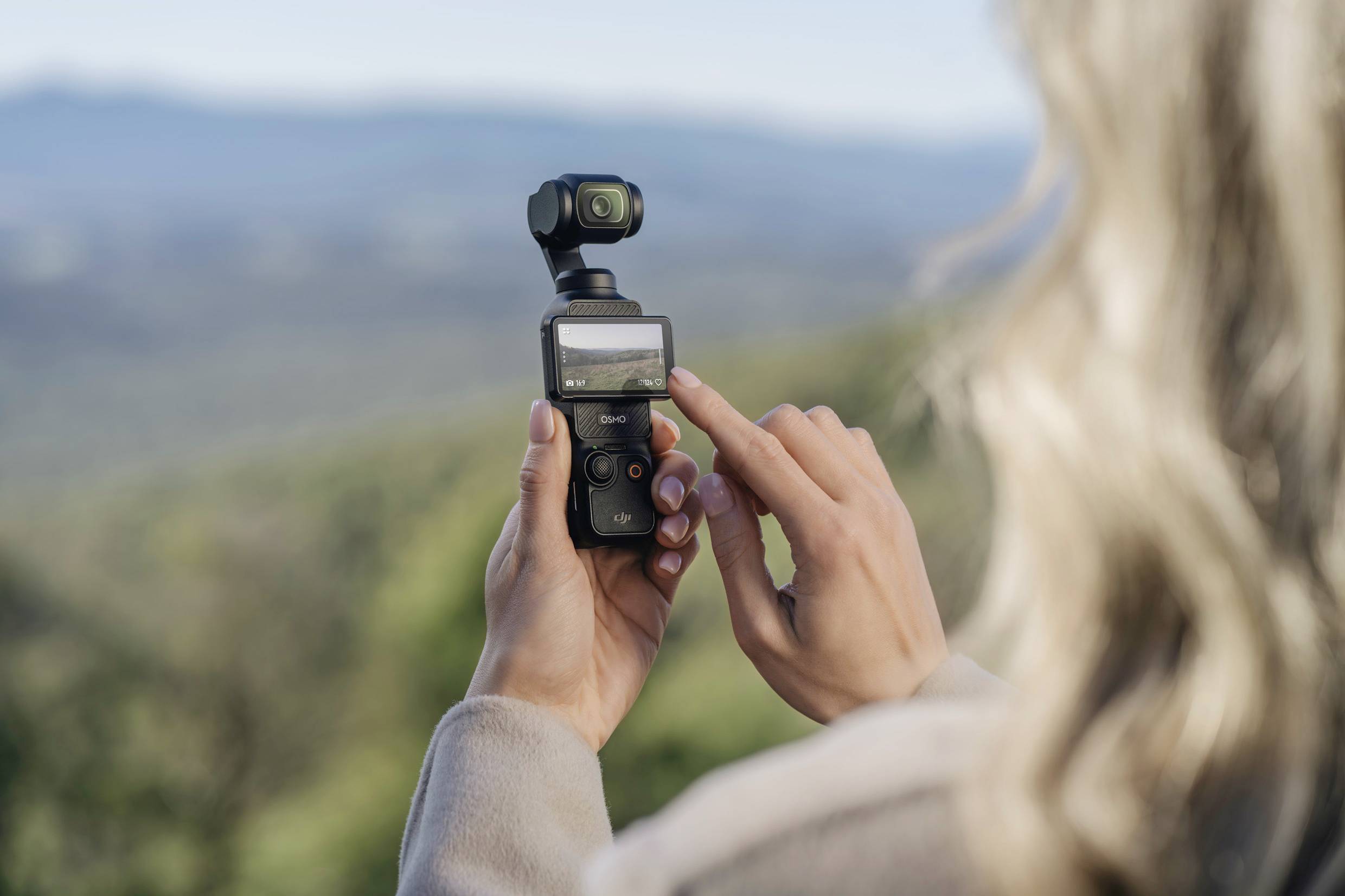 A person is holding a small camera with a display showing a landscape. The camera is pointing towards a wooded environment.