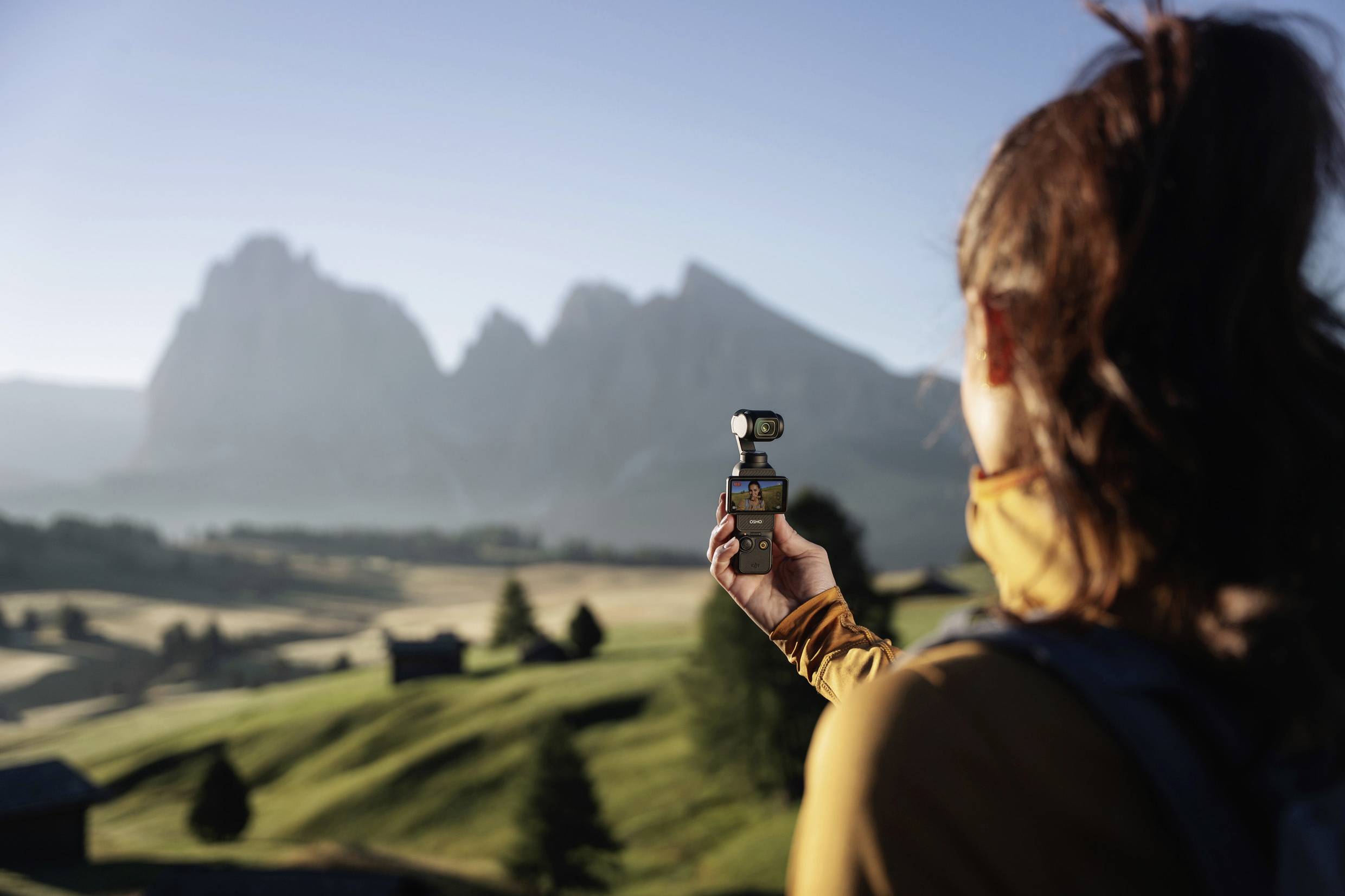 A woman is holding a camera and filming a mountain landscape in the early morning hours. Hills and mountains can be seen in the background.