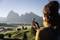 A woman is holding a camera and filming a mountain landscape in the early morning hours. Hills and mountains can be seen in the background.