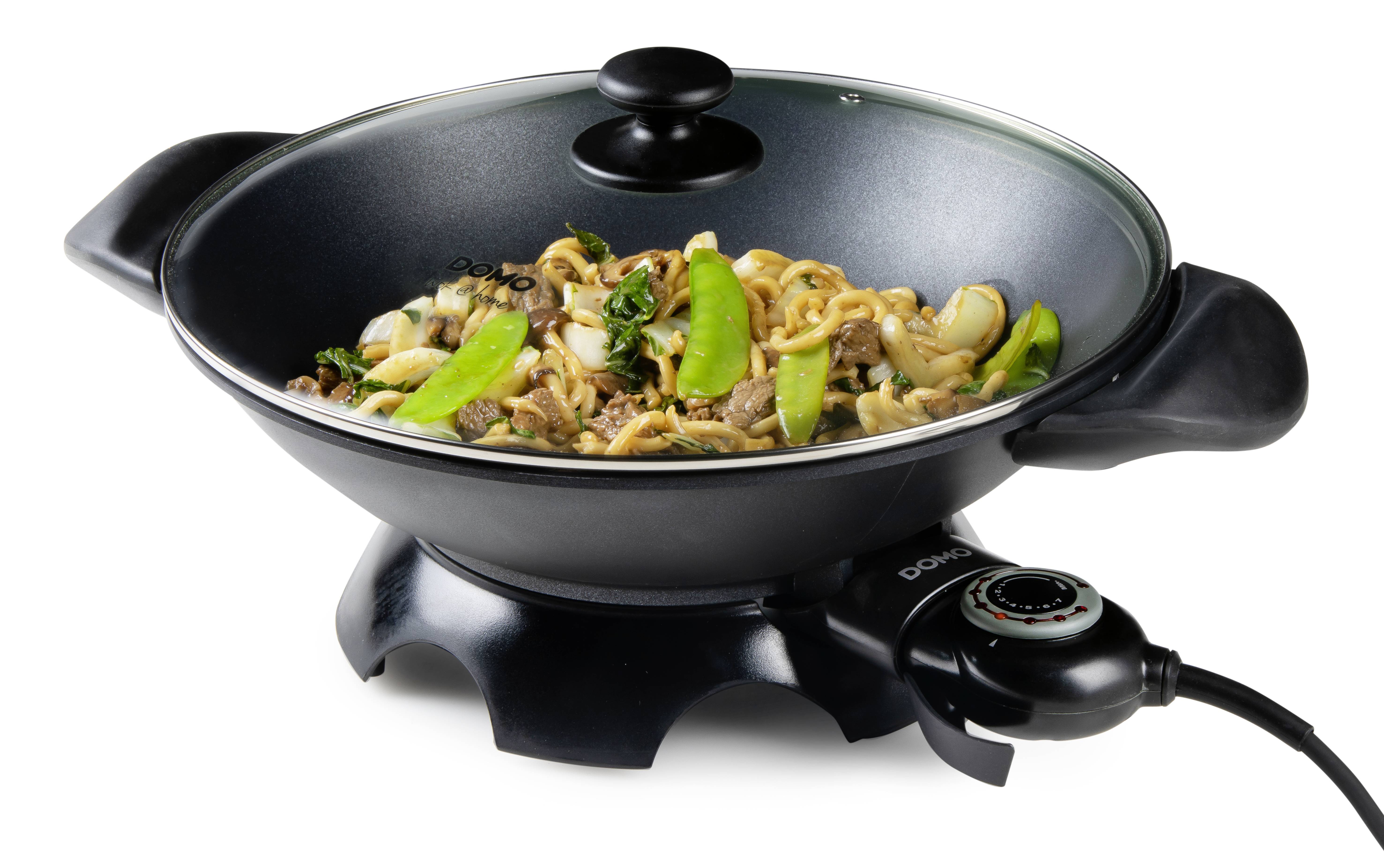 An electric wok filled with vegetables and noodles, with identifiable green sugar snap peas and brown mushrooms, placed on a white surface.