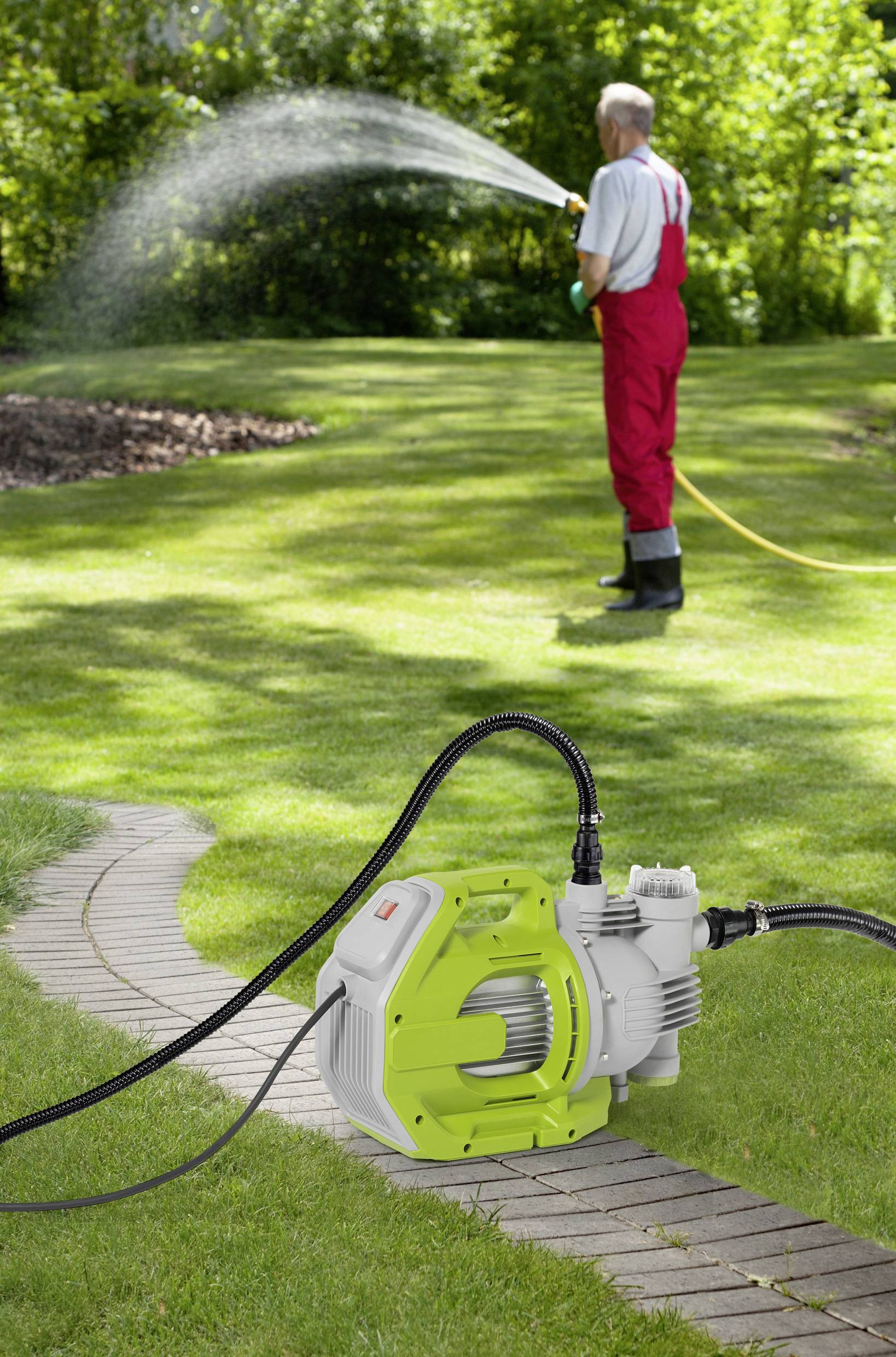 A man in a red overall is watering a lawn with a garden hose. In the foreground, a green electric water pump stands on a path.
