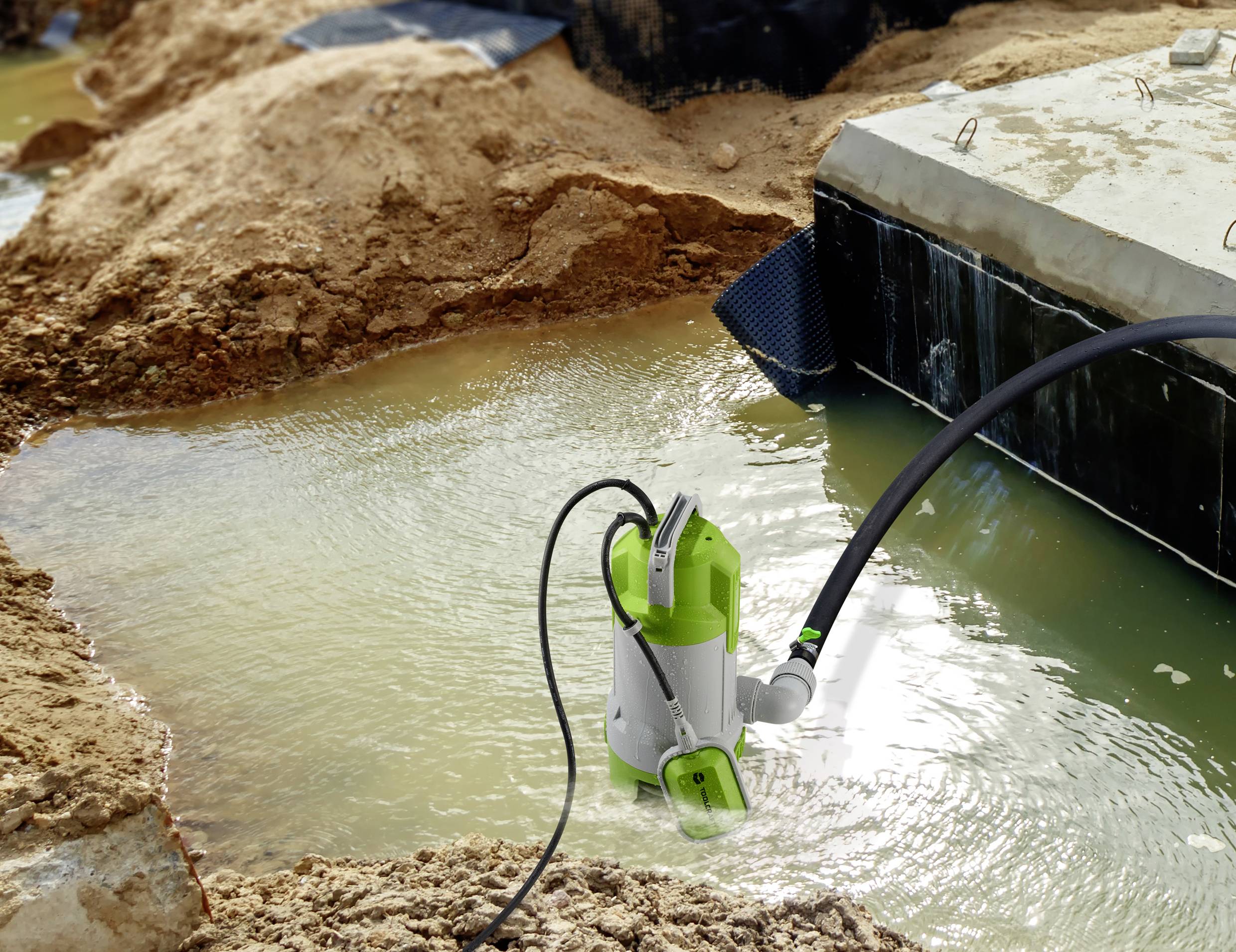 A green water pump is draining a pit on a construction site. The surrounding area is muddy, and water is being pumped out.
