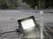 A glowing LED floodlight lies in the rain on a wet cobblestone street, water splashing around it.