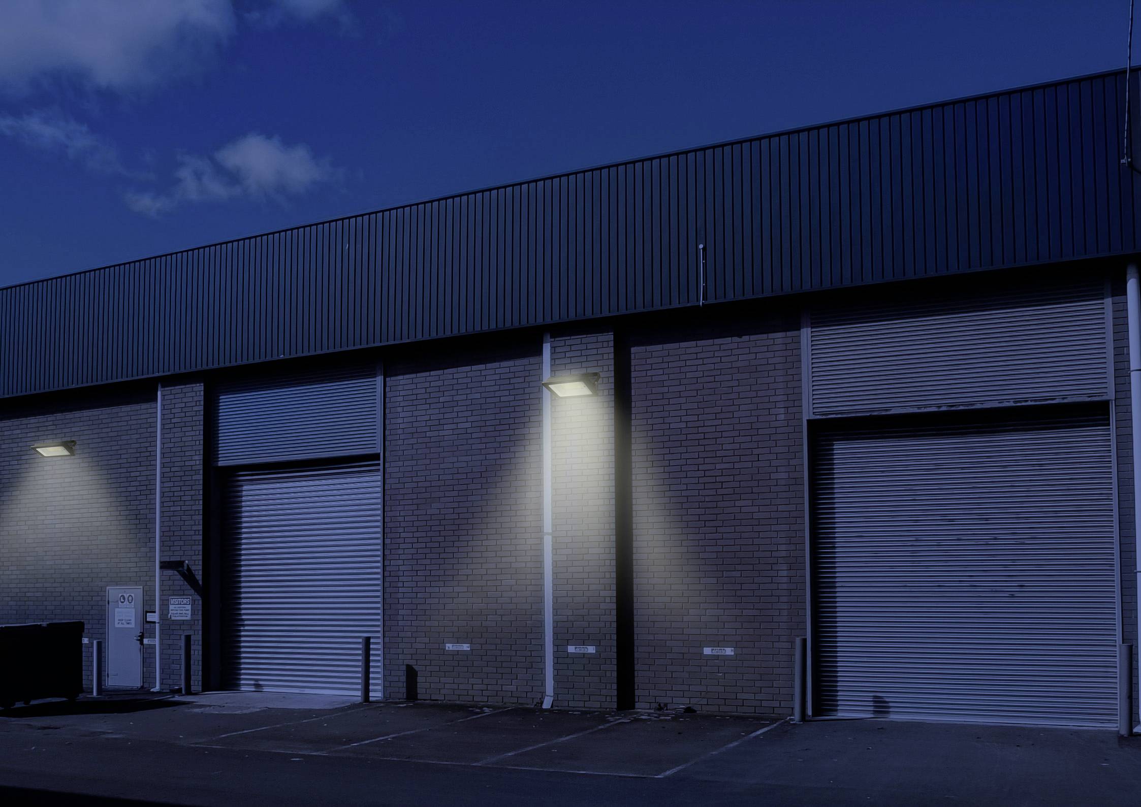 Industrial buildings at night with illuminated garage roller shutters, concrete walls, and a partly cloudy sky in the background.