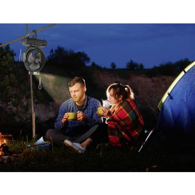 A man and a woman are sitting next to a tent at night, drinking from mugs and kept warm by a blanket, beneath a hanging fan.