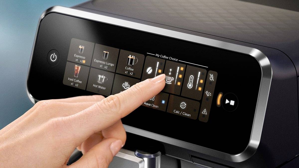 A hand selects a coffee option on the touchscreen of a coffee machine. Various coffee beverages are visible.