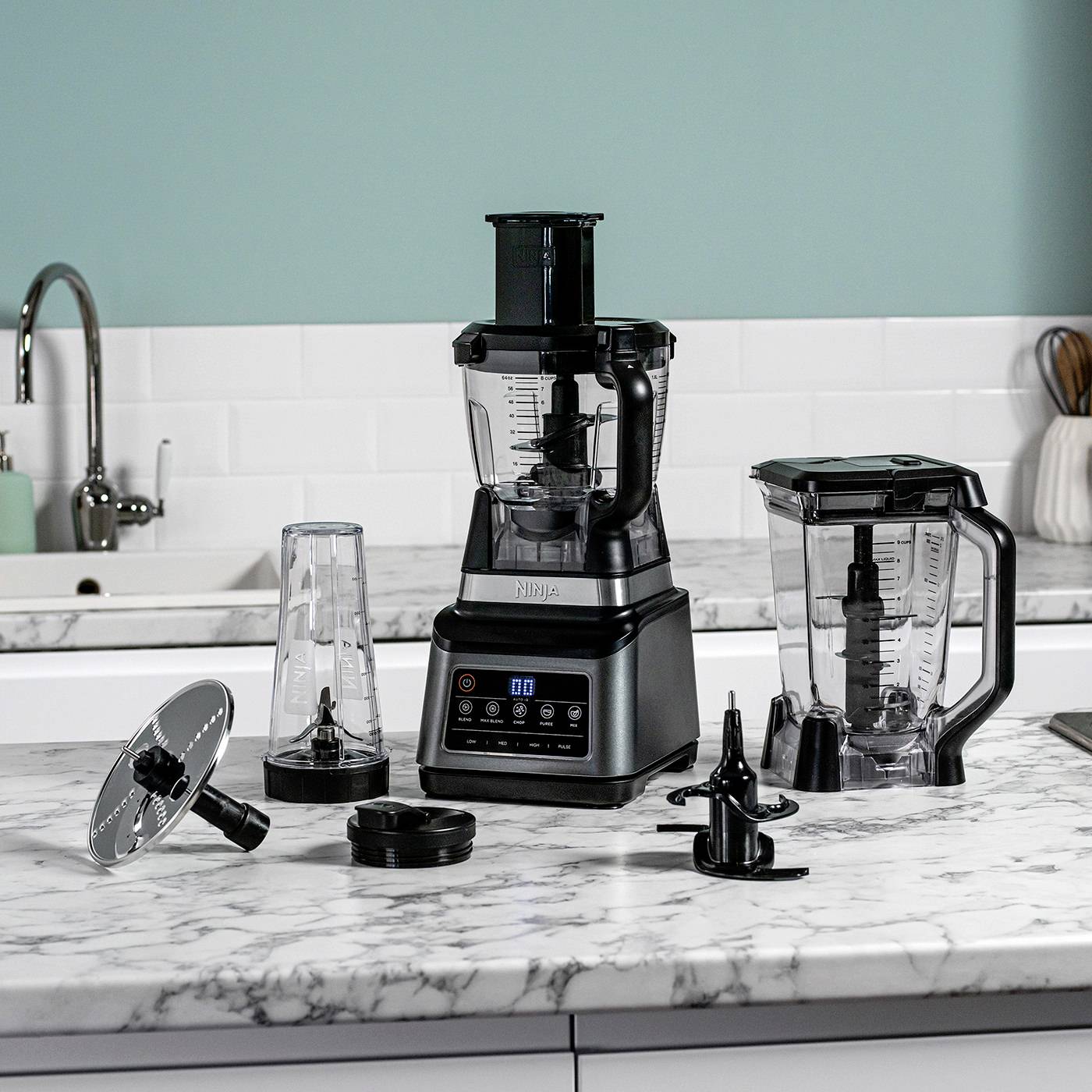 A black mixer with several interchangeable attachments sits on a marble kitchen worktop. A sink is visible in the background.