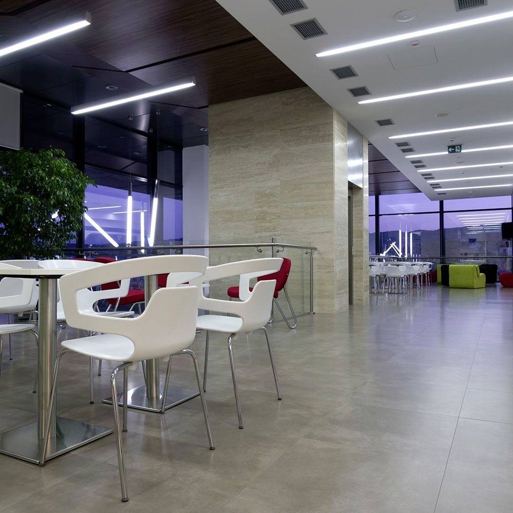 A modern break room with white chairs and tables, large windows and bright lighting. A plant stands in the corner.