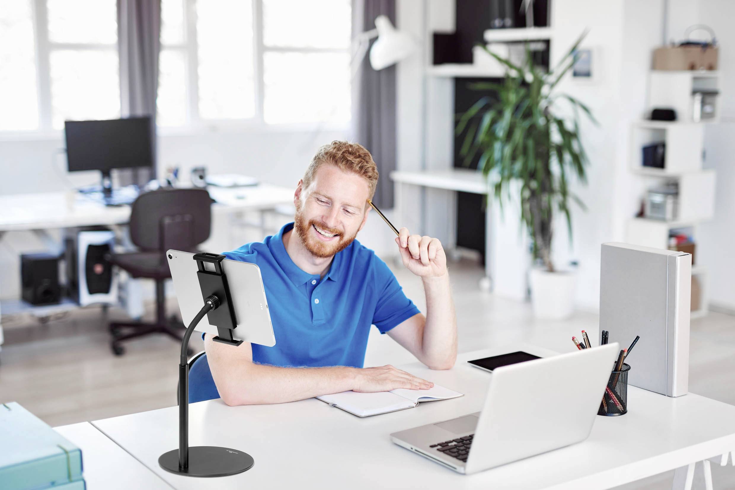 A man is sitting at a desk in a modern office, smiling and holding a pen. In front of him are a laptop and a tablet.