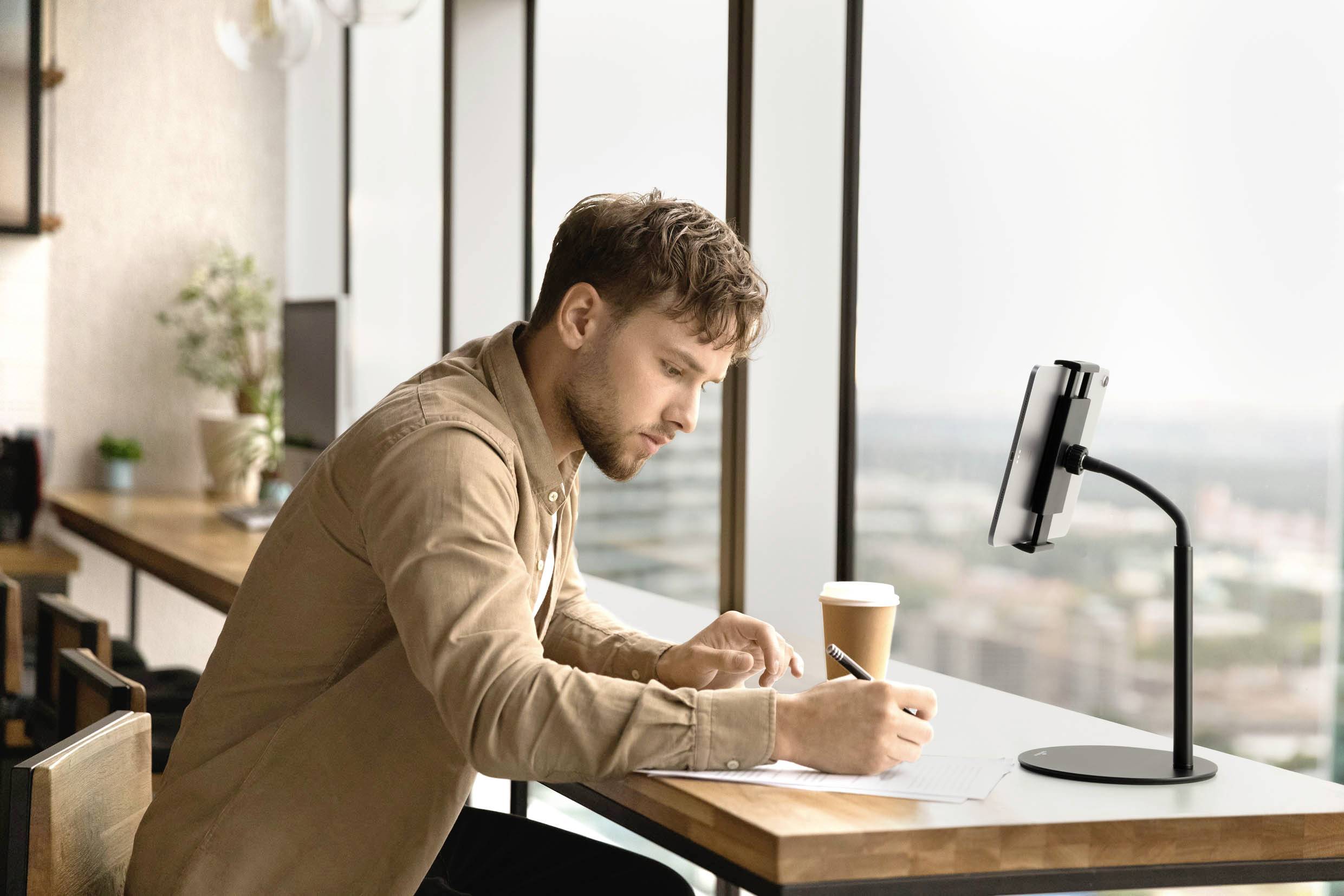 A man is sitting at a table, writing on paper and holding a disposable cup. A smartphone is positioned on a stand beside him.