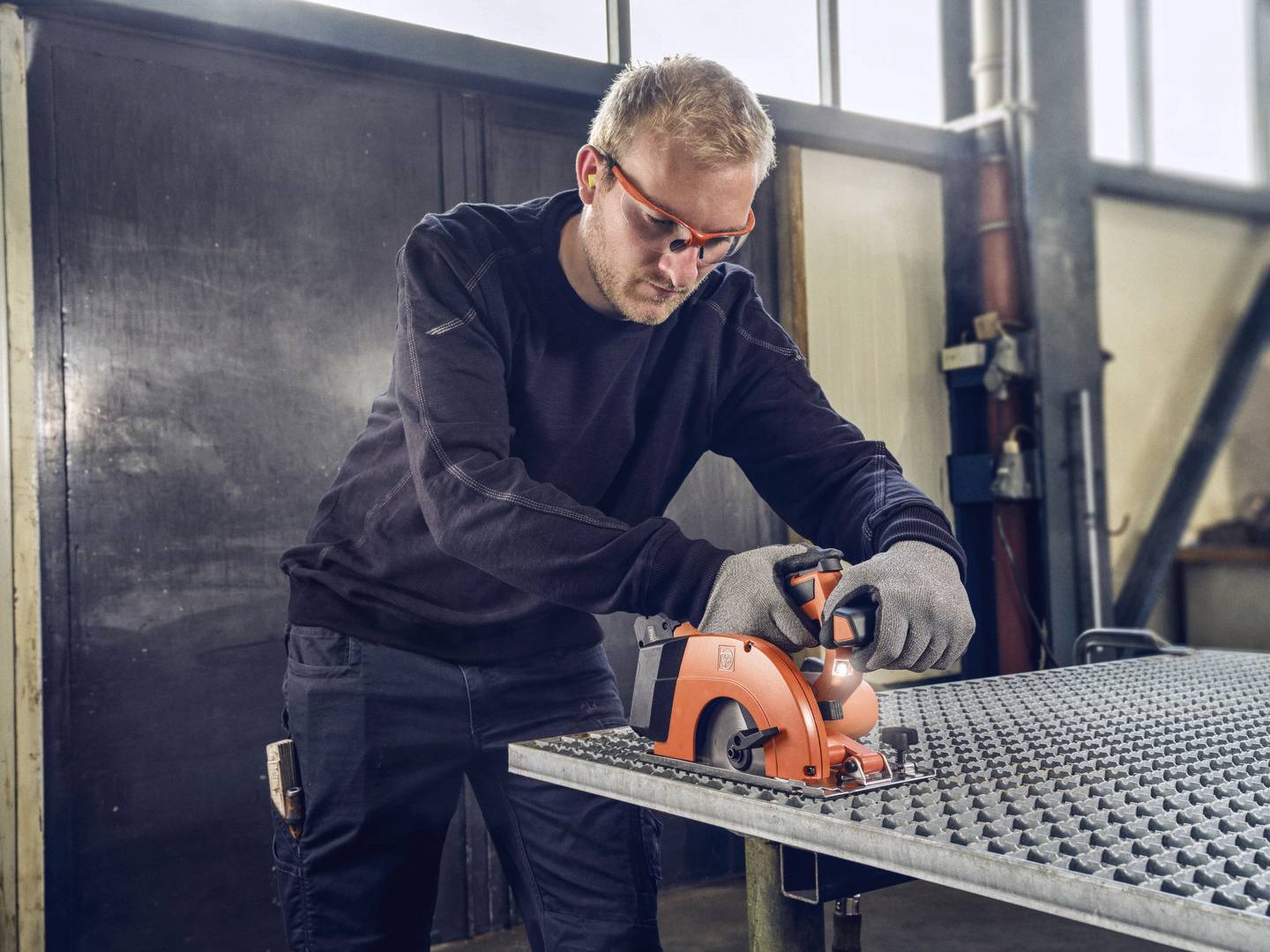 A man wearing safety glasses and gloves is using a handheld circular saw to cut metal on a table in a workshop.