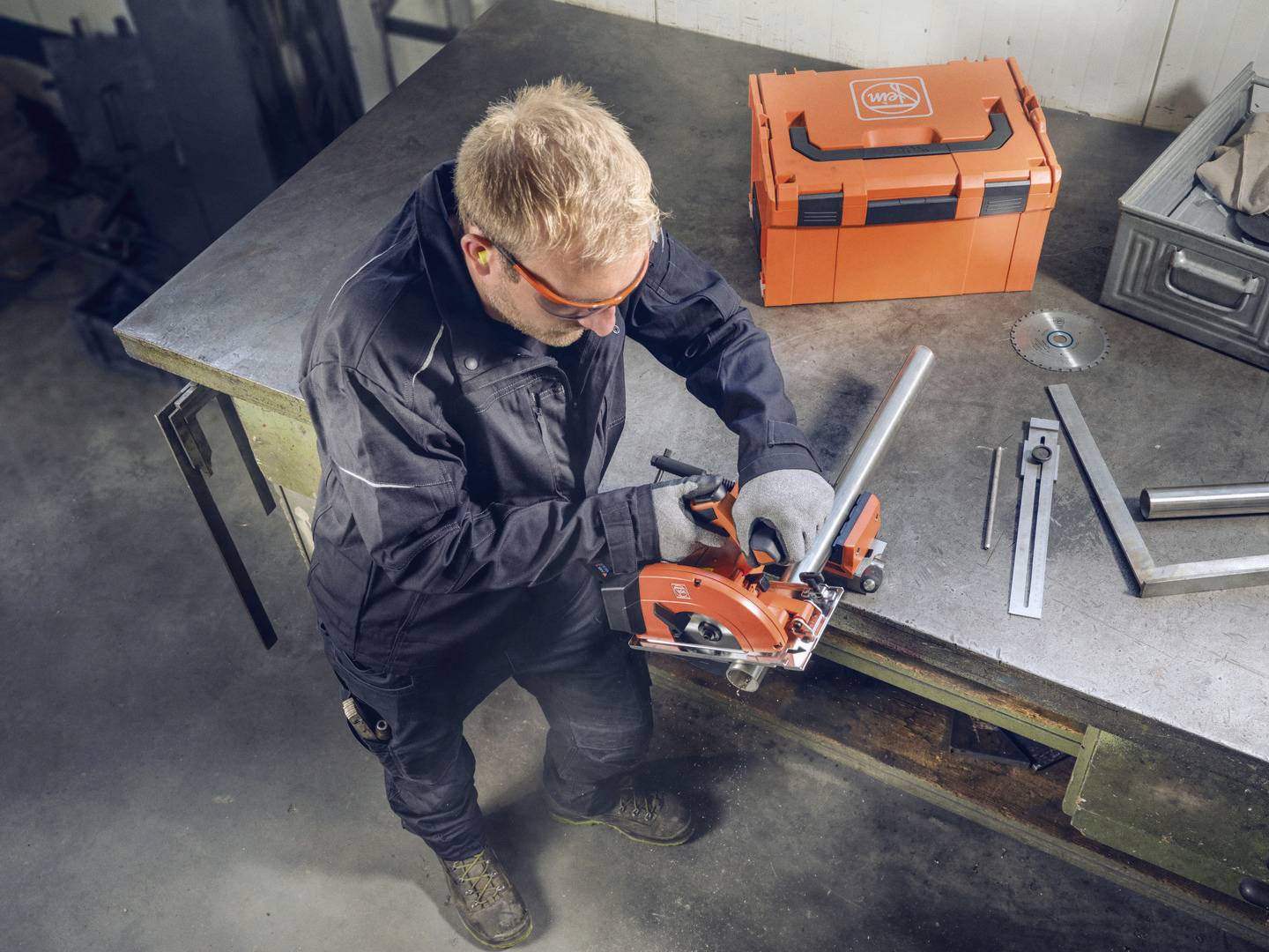 A man in workwear is cutting a metal pipe with an orange saw on a workbench. Tools are scattered in the background.