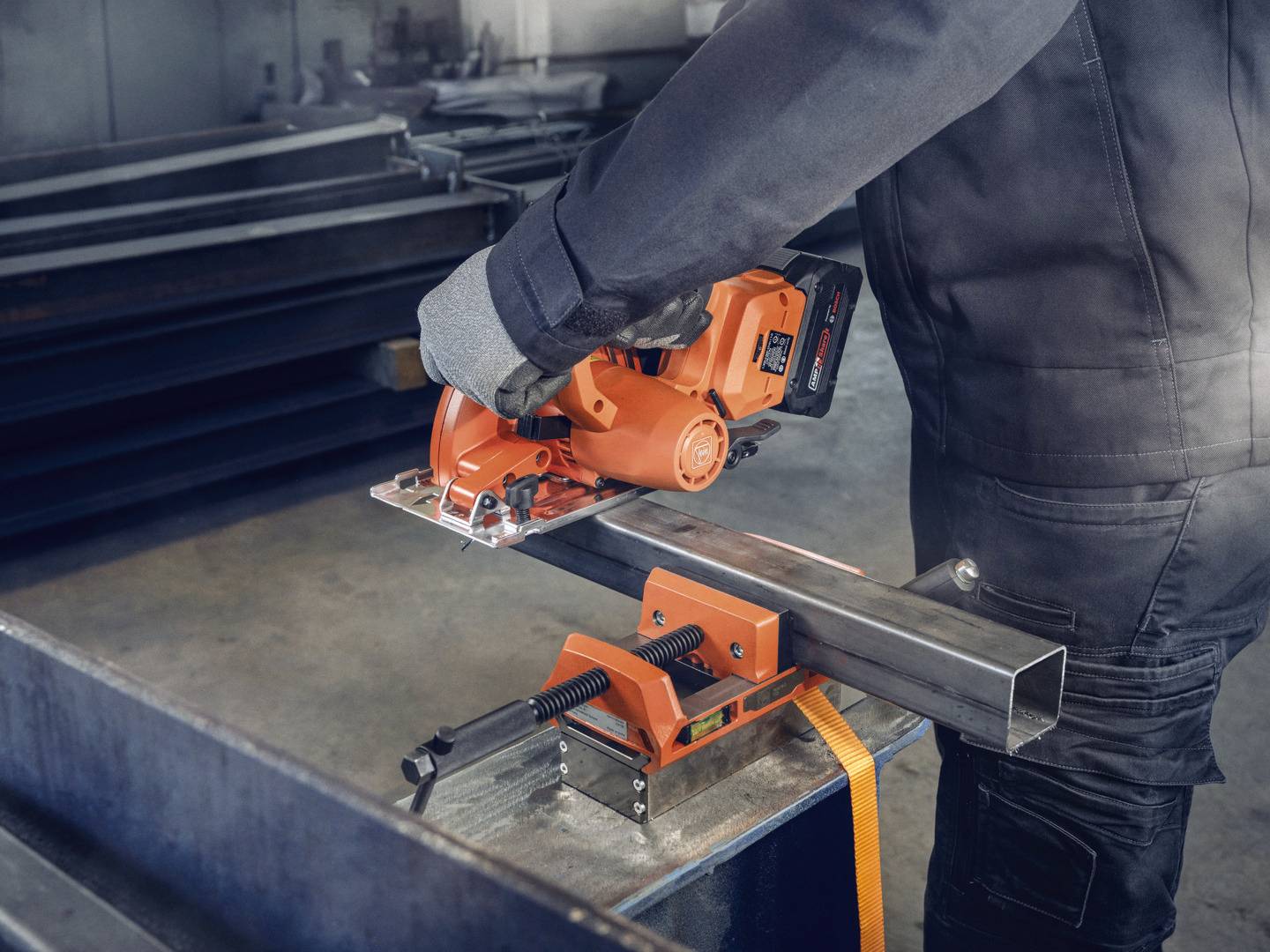 A person is working with an orange, portable metal saw to cut a square metal tube. Protective clothing is being worn.