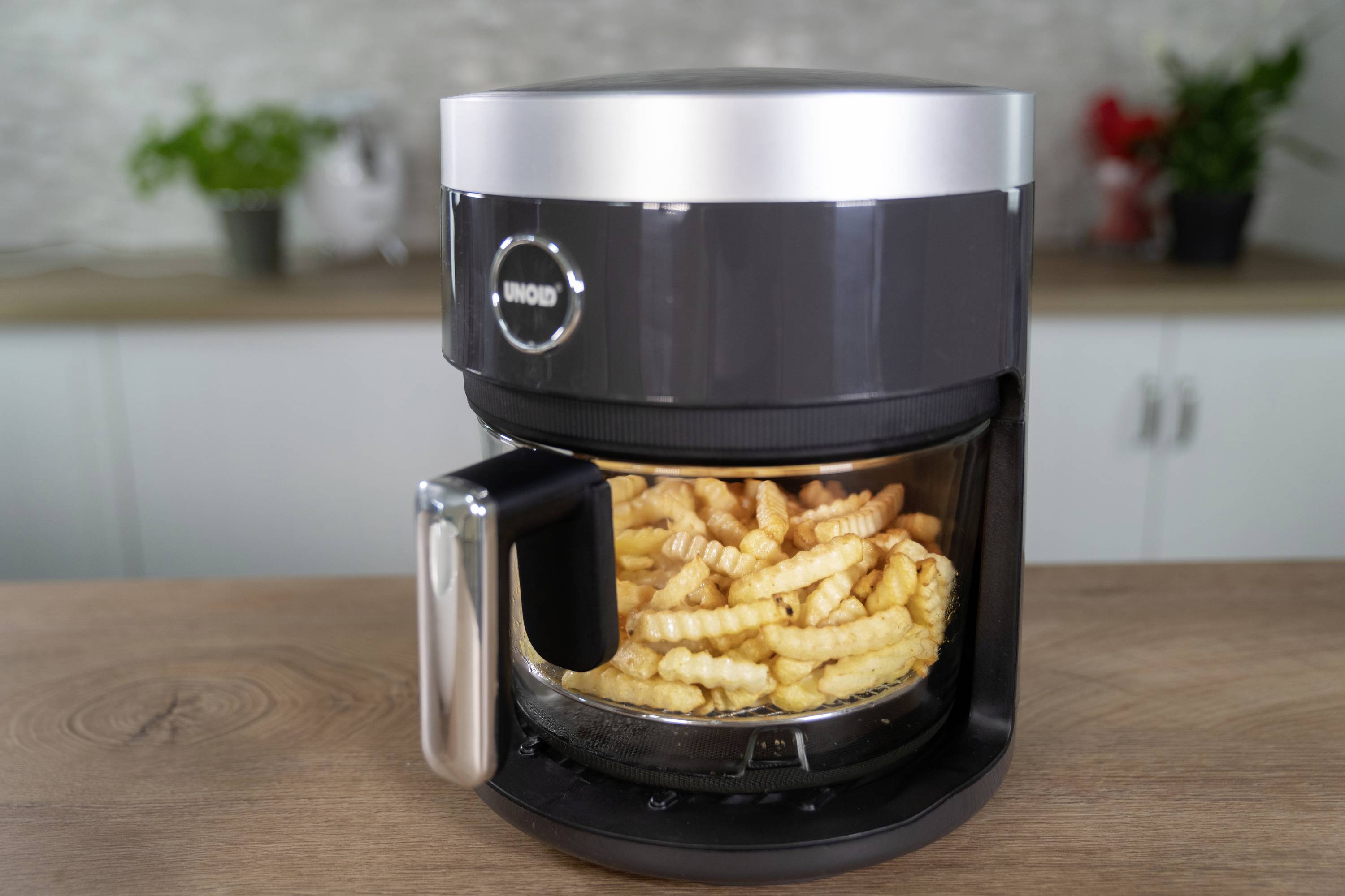 A black air fryer sits on a wooden worktop. Wavy chips can be seen inside. A kitchen is visible in the background.