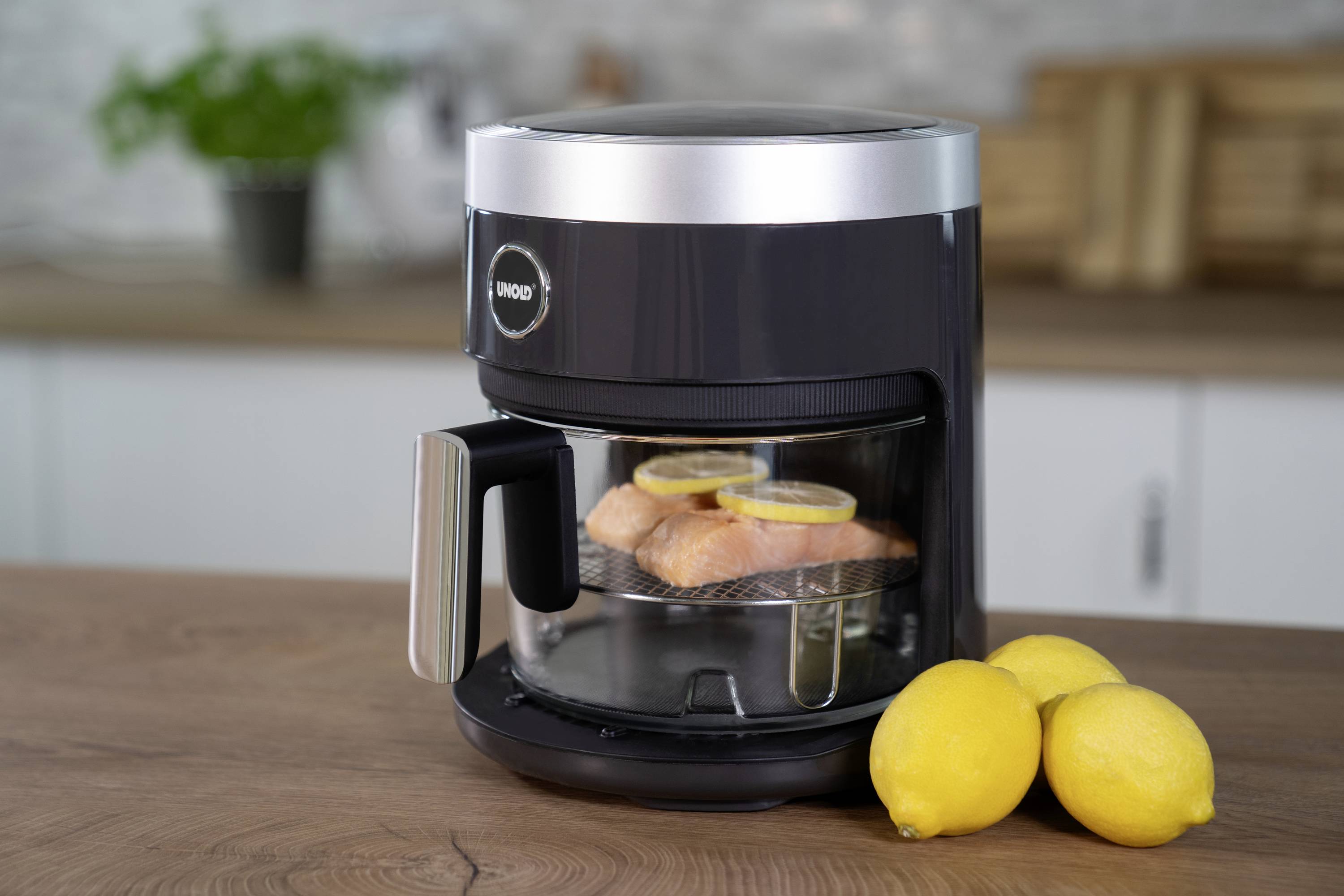 A black air fryer with a window sits on a kitchen worktop. Chicken thighs are sizzling inside with lemon slices.