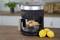 A black air fryer with a window sits on a kitchen worktop. Chicken thighs are sizzling inside with lemon slices.