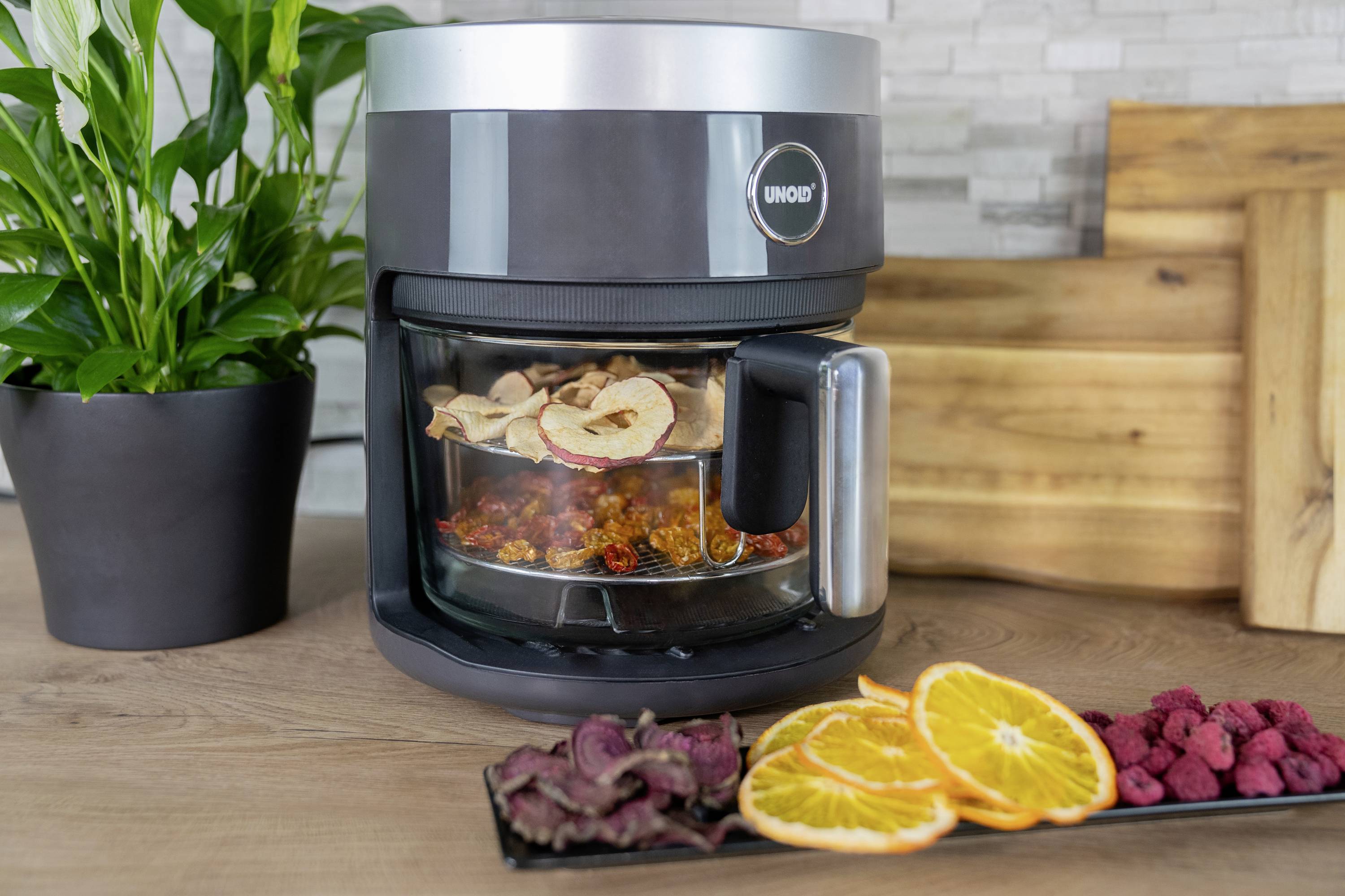 A food dehydrator with dried fruits and vegetables on a tray. In the background, there are chopping boards and a plant.