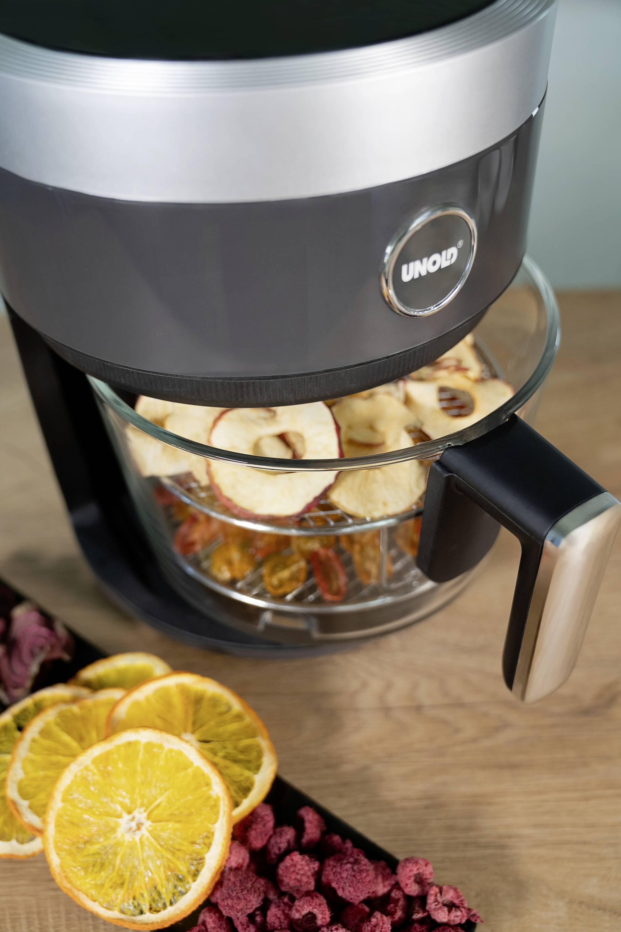 A dehydrator dries apple slices, oranges and berries. It stands on a wooden table next to fresh, sliced oranges.