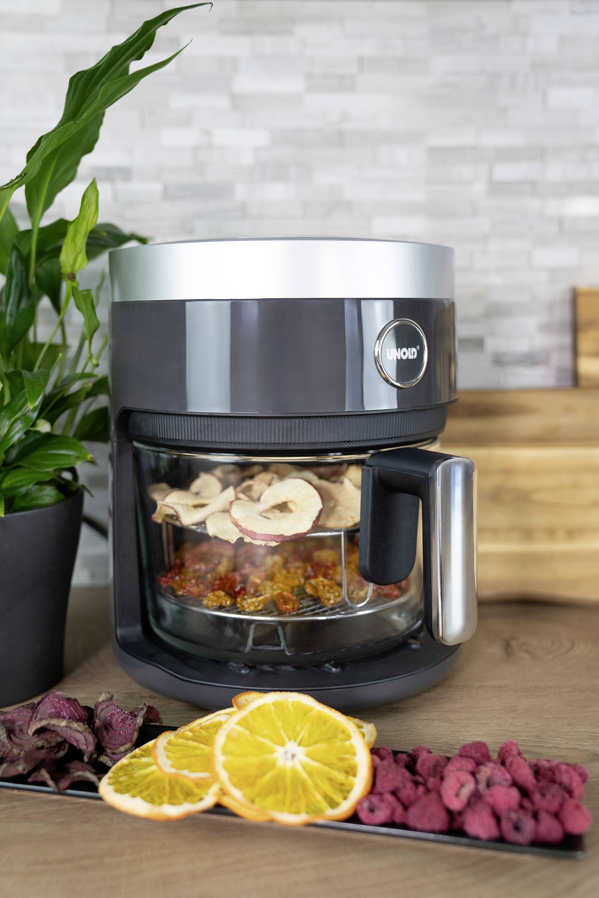 A modern dehydrator on a kitchen worktop with dried apple and orange slices in a glass container, with berries alongside.