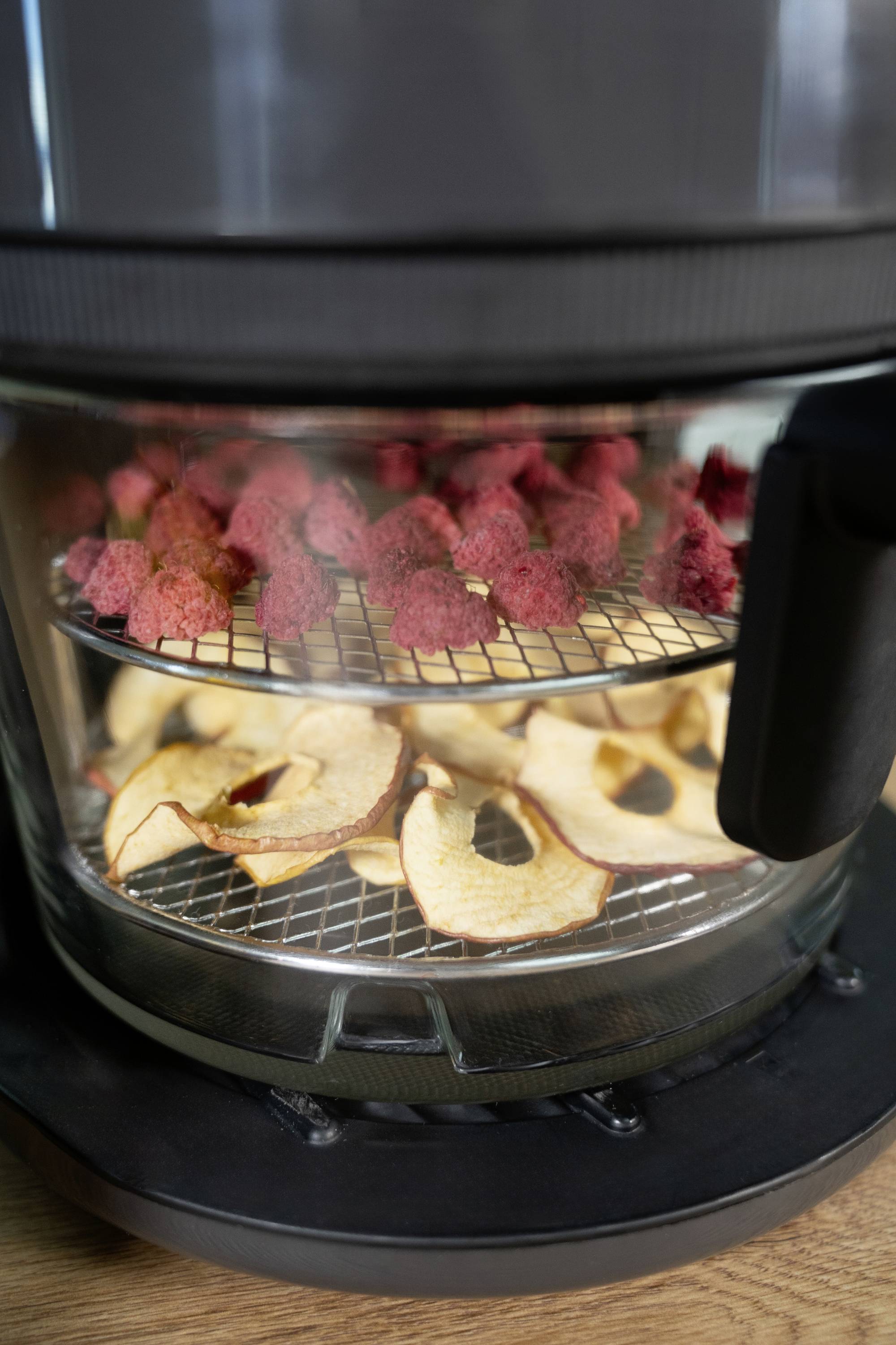 A two-tier food dehydrator with dried fruits: red fruits on the top shelf, apple slices on the bottom shelf. The device is positioned on a wooden table.