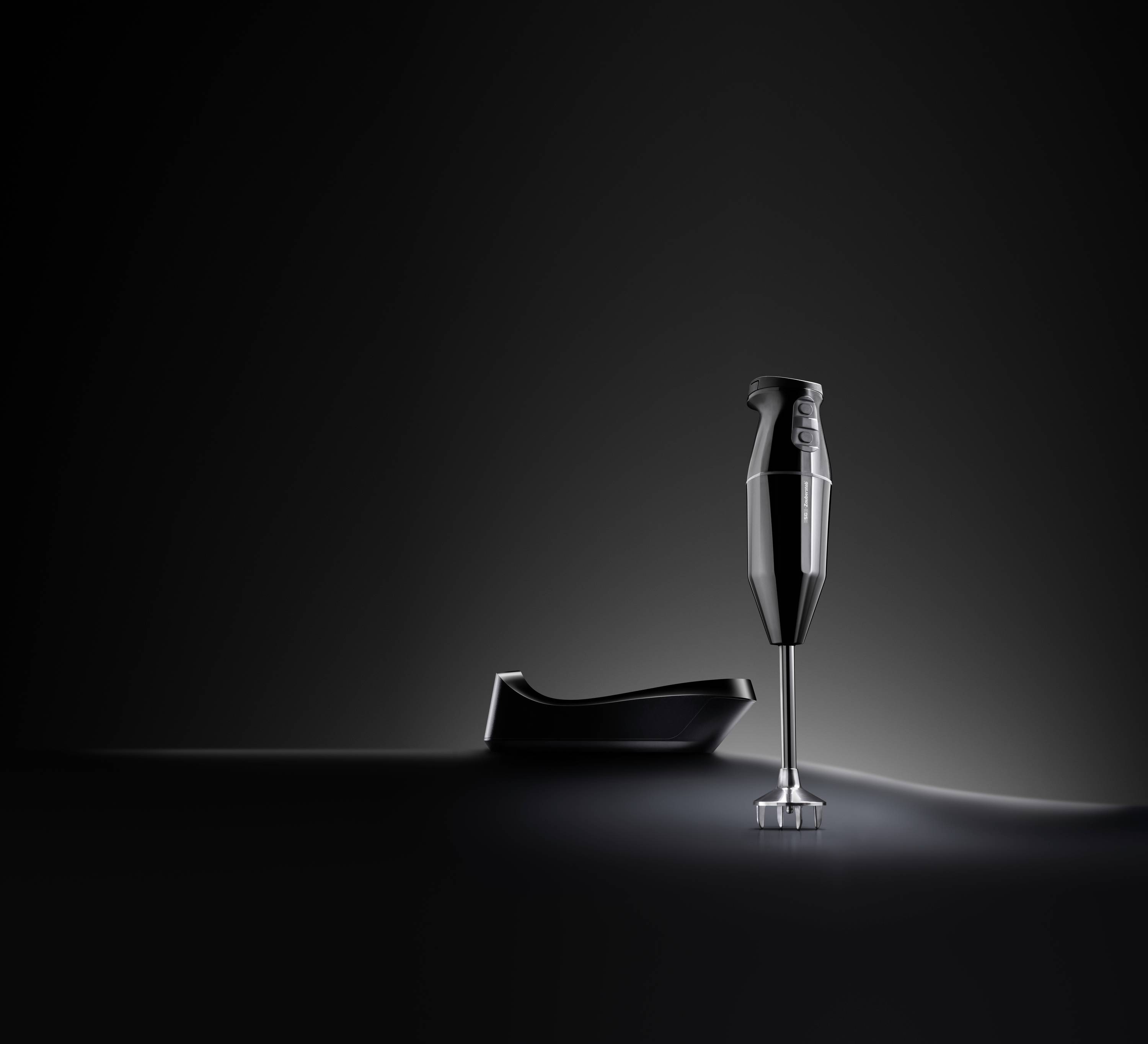 A black hand blender stands upright next to its black holder against a dark background.