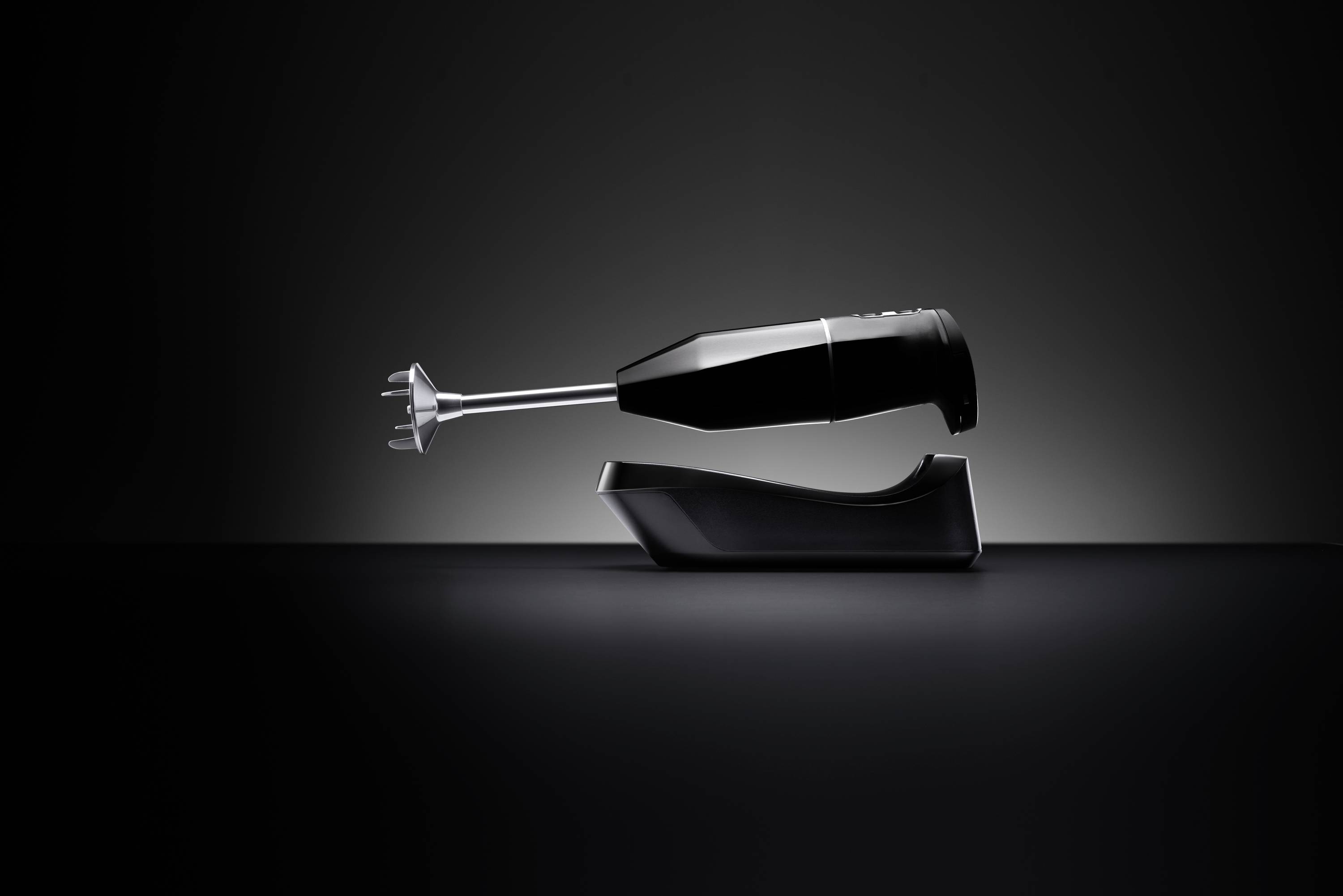 A black hand blender with silver blades rests on a black stand. The background is dark and minimalist.