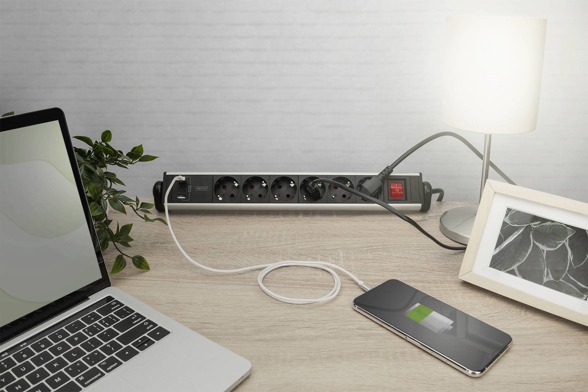 A desk with a laptop, a charger charging a smartphone, a desk lamp, and a framed picture of a plant.