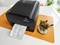 A thermal printer on a table is printing a receipt. Next to it, plants are sitting on an orange placemat.